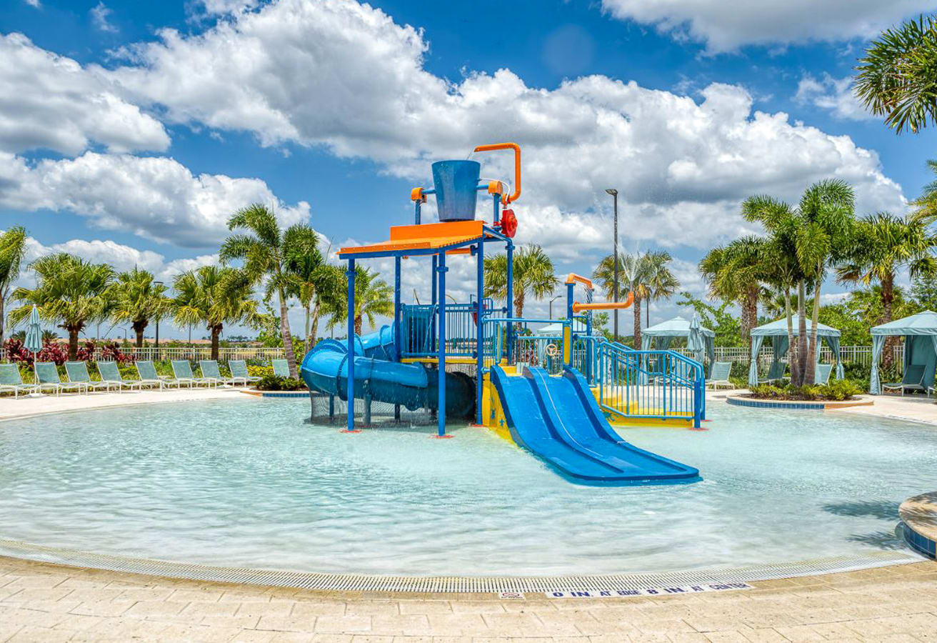 A shallow, zero-entry pool area designed for children, featuring a colorful blue and yellow water play structure with slides, a dumping bucket, and spray features under a partly cloudy sky.