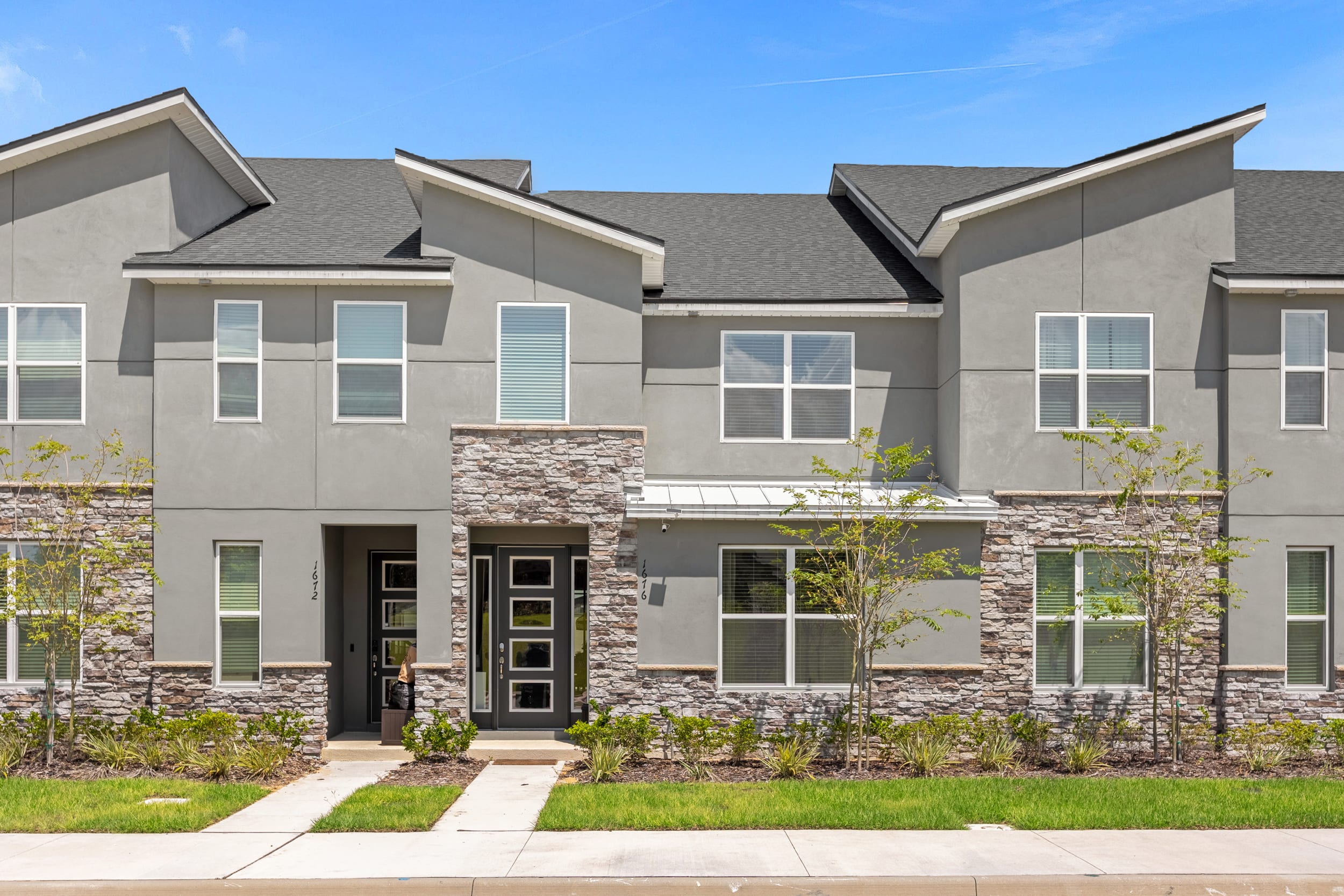 The front exterior of a modern townhouse, featuring a gray stucco facade with stone accents on the lower level and around the entrance. Multiple windows with blinds are visible, set against a clear blue sky.