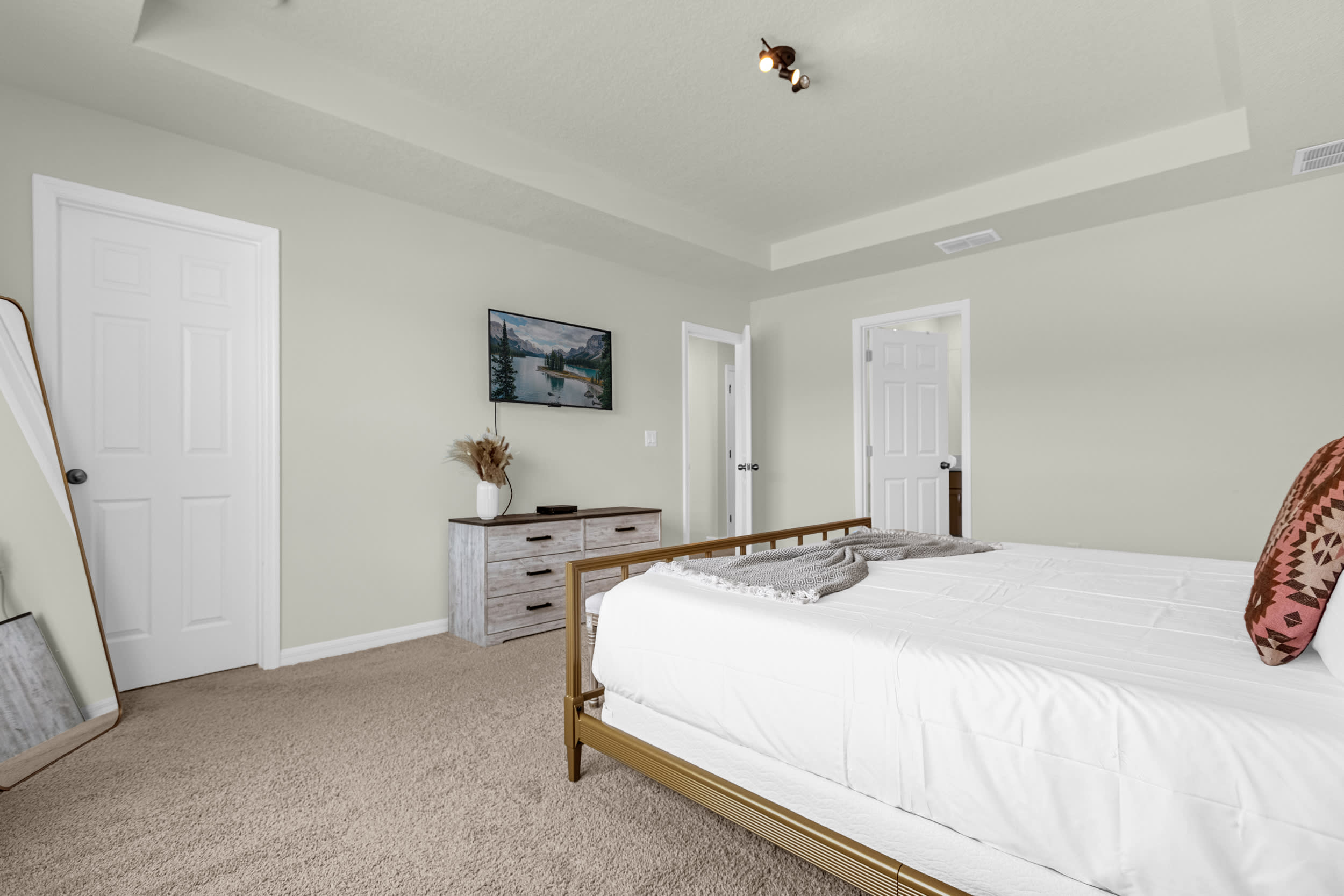 View of the primary bedroom showing a wall-mounted TV above a rustic dresser, a full-length mirror, and a white door, with a glimpse of the gold metal bed frame.