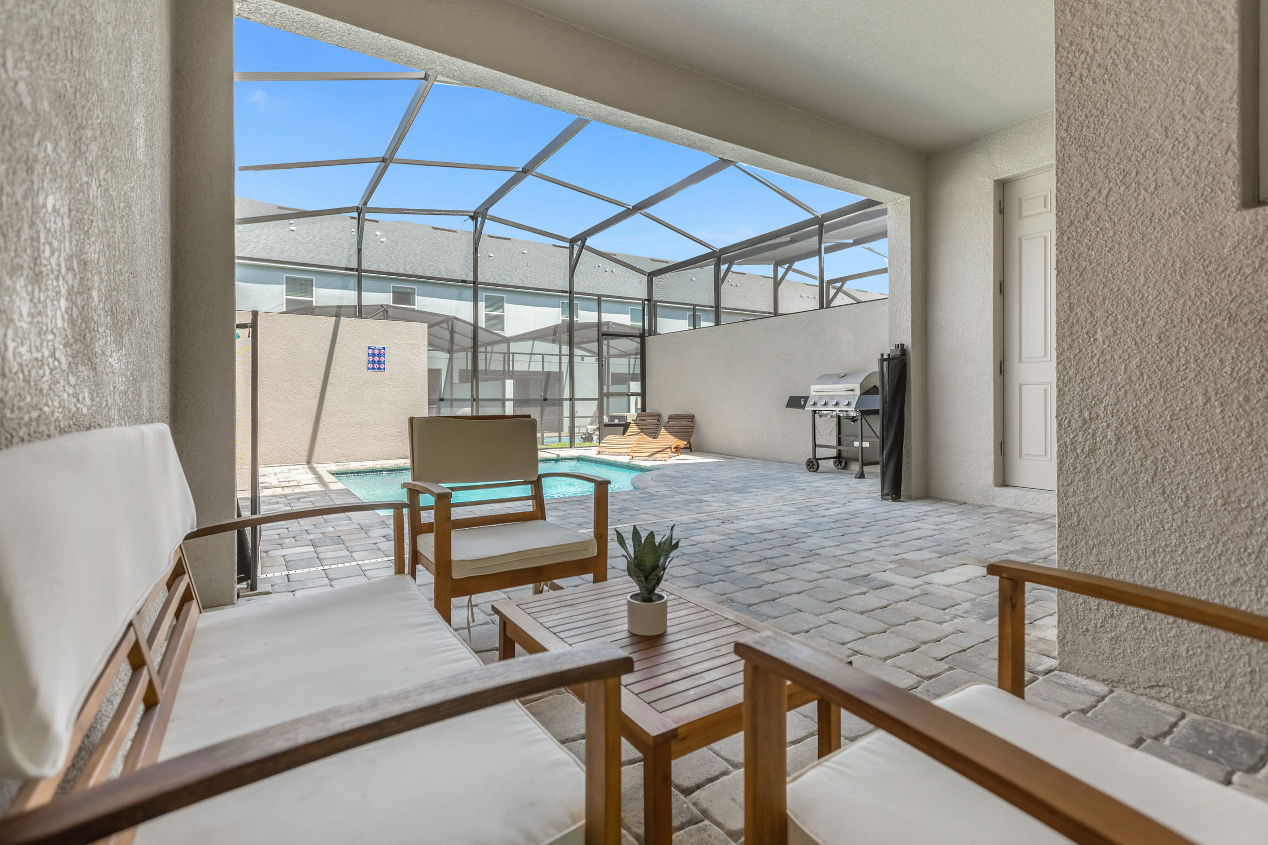 Covered outdoor patio area with a set of wooden chairs and a coffee table, offering a comfortable seating space. The area overlooks the screened private pool and features paver flooring.