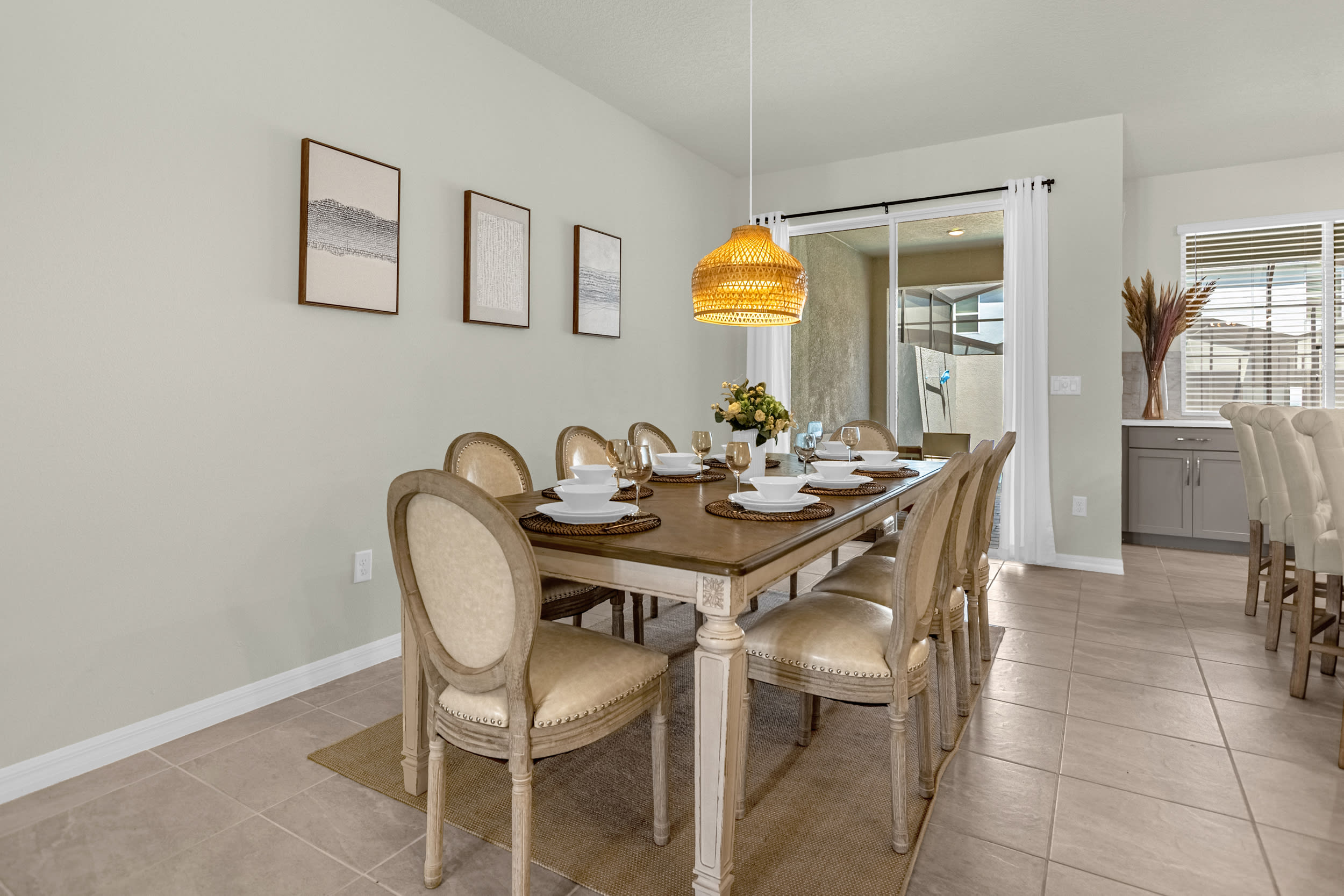 Elegant dining room featuring a long wooden table set for eight, surrounded by upholstered chairs with light frames. A woven pendant light hangs above the table, and a sliding glass door leads to an outdoor patio.