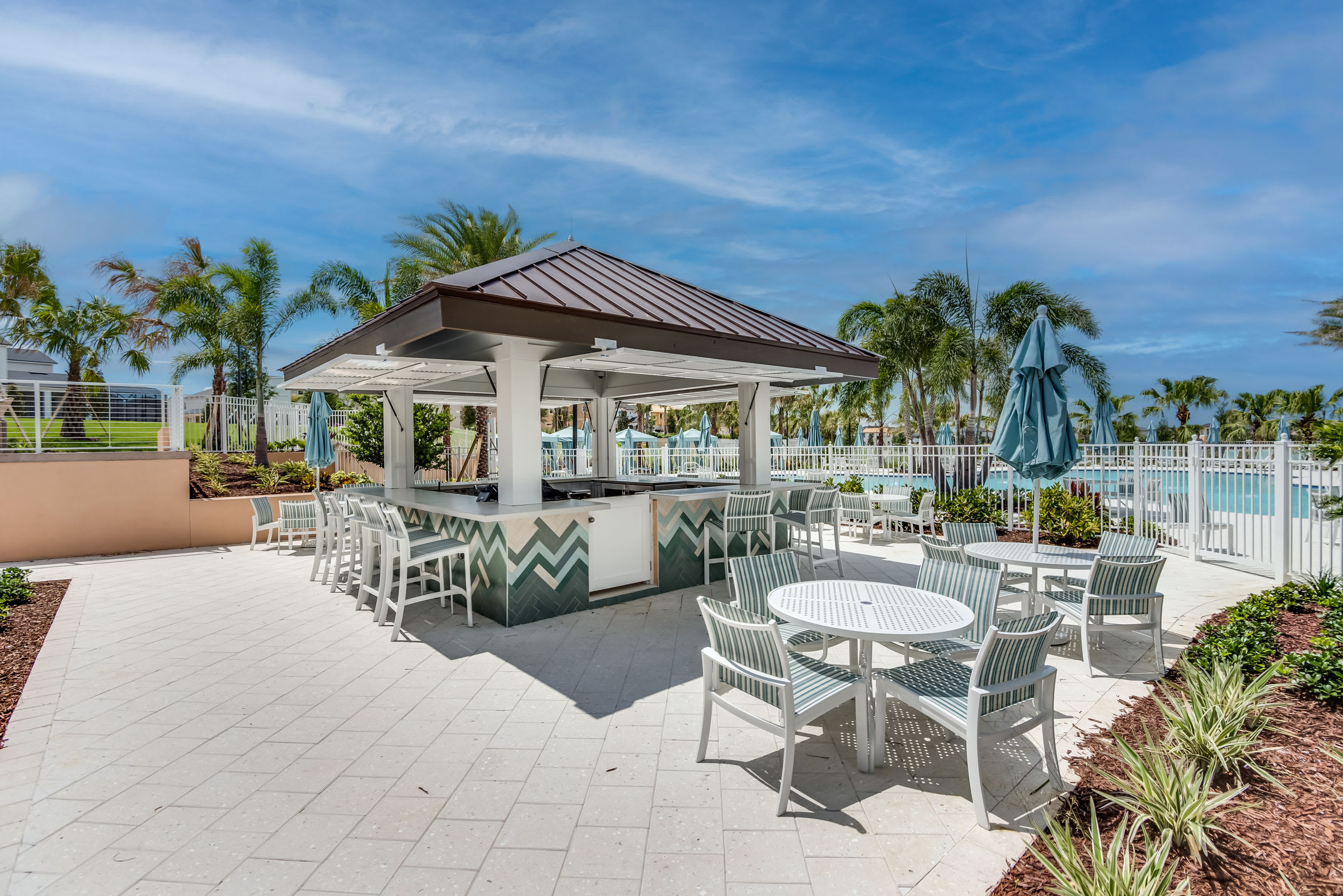 An inviting outdoor bar area with a covered counter, bar stools, and several round tables with chairs, situated on a paved patio adjacent to a communal swimming pool under a bright blue sky.