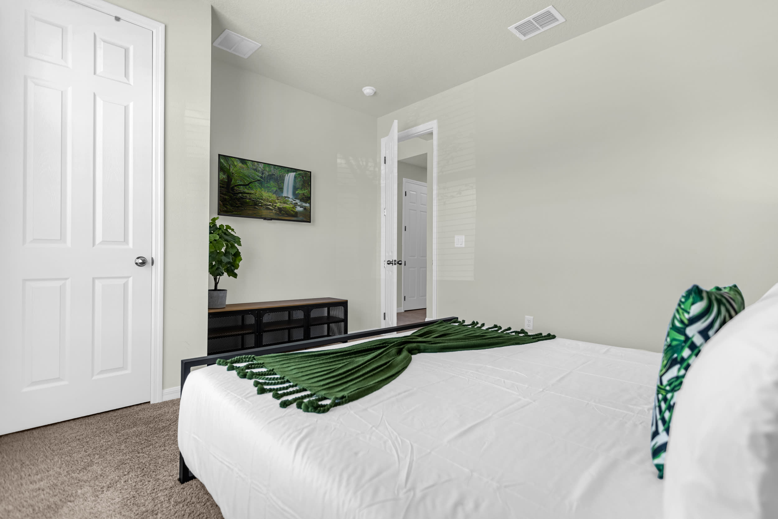 Another view of the guest bedroom showing a wall-mounted TV above a dark wood console, a potted plant, and a white door, with a green throw blanket on the bed.