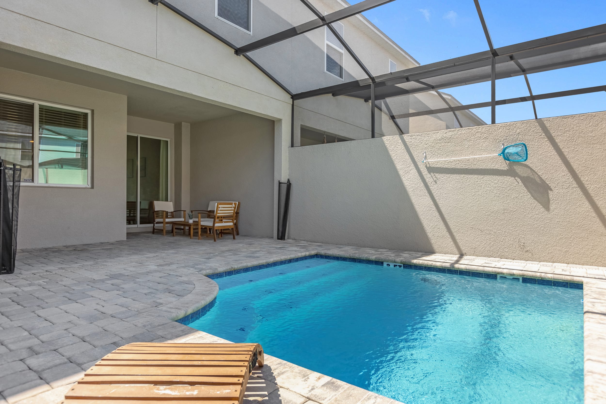 View of the private screened pool area, showing the pool with blue water, a wooden lounge chair, and a paved patio. The surrounding walls and screen enclosure provide privacy, with a glimpse of the house exterior.