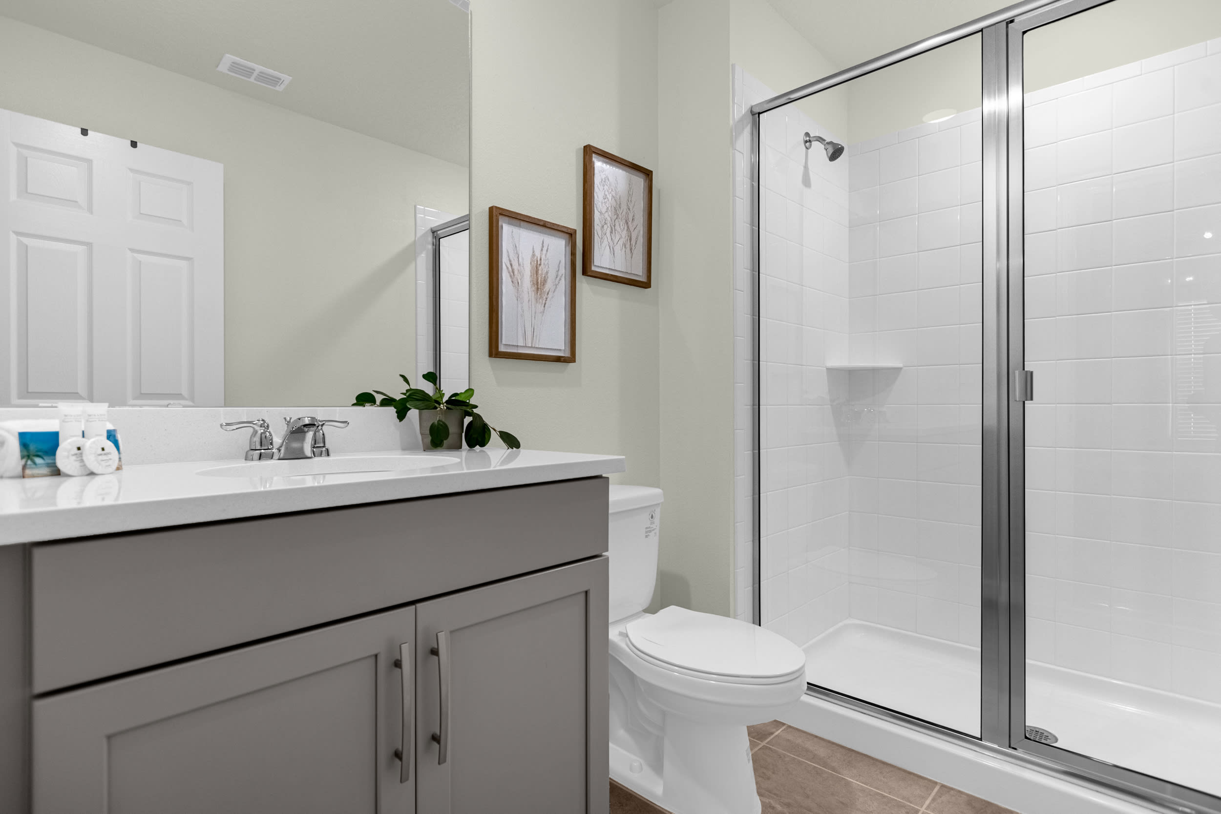 A modern bathroom with a light gray vanity, white quartz countertop, and a large mirror. It features a walk-in shower with white subway tiles and a glass enclosure, alongside a white toilet.