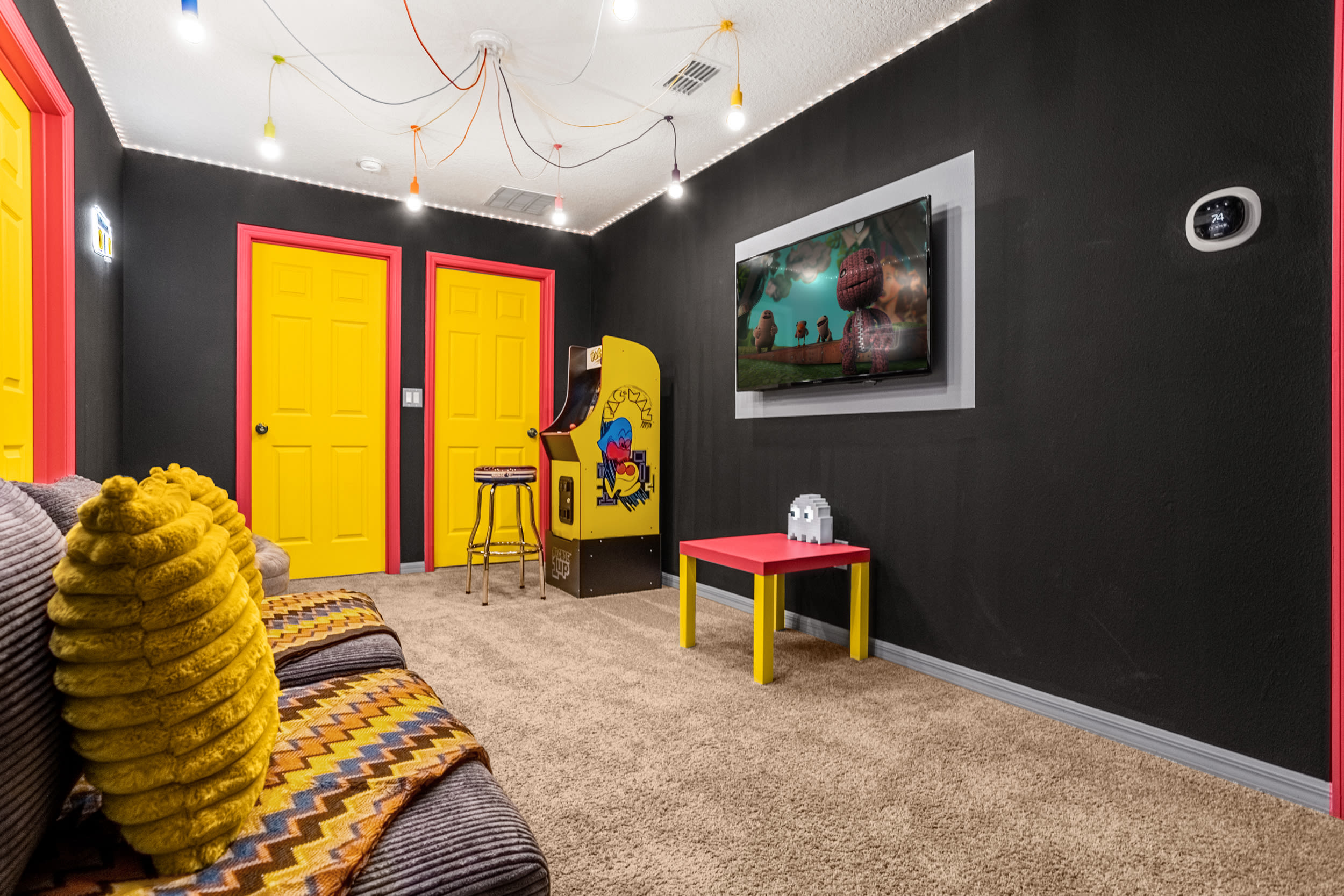 Another view of the vibrant game room, showcasing a wall-mounted TV, a yellow arcade machine, and a small red table with a pixelated ghost decoration. The room features dark walls, yellow doors, and string lights.