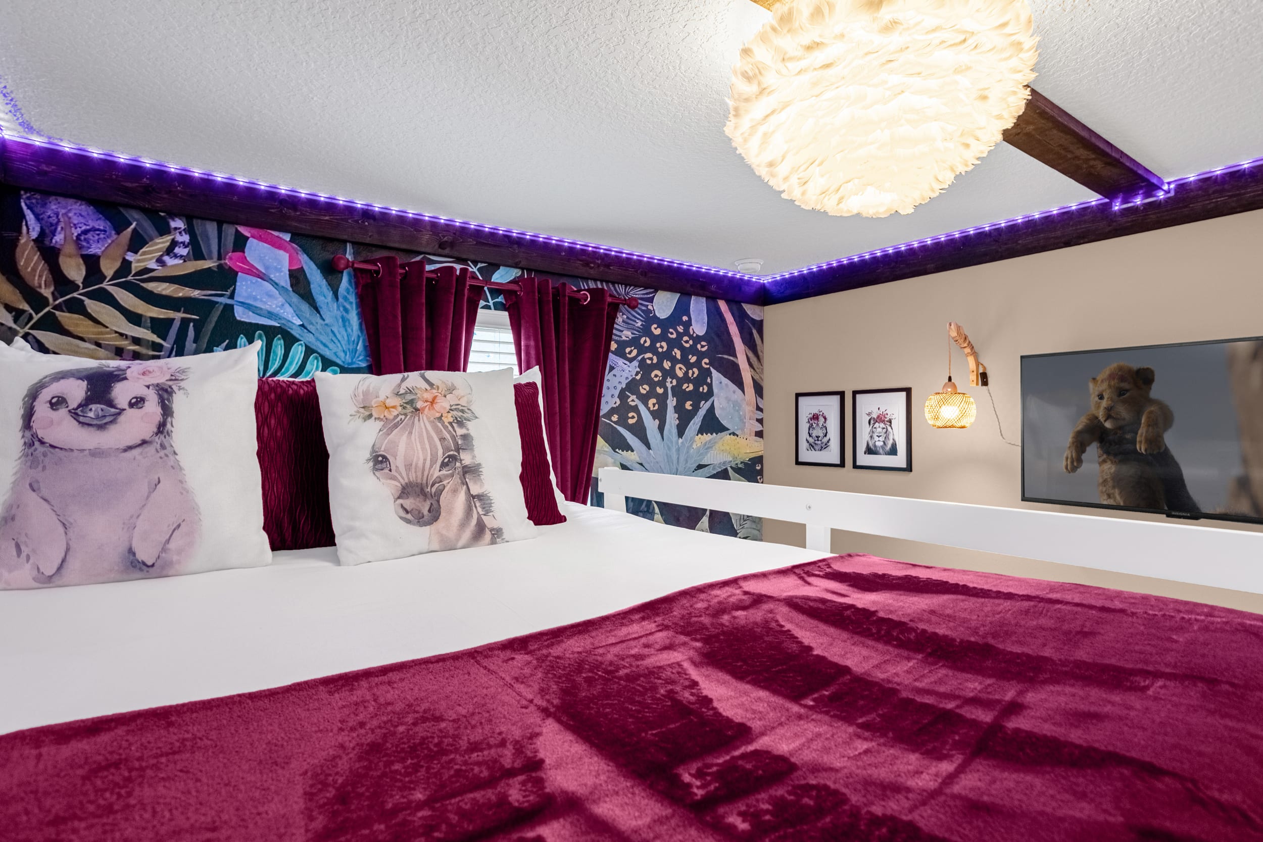 A vibrant themed bedroom featuring a bunk bed with a white frame and a deep red comforter, accented by animal-themed pillows. The room has a dark floral accent wall, purple LED strip lighting, and a TV showing an animated lion cub.