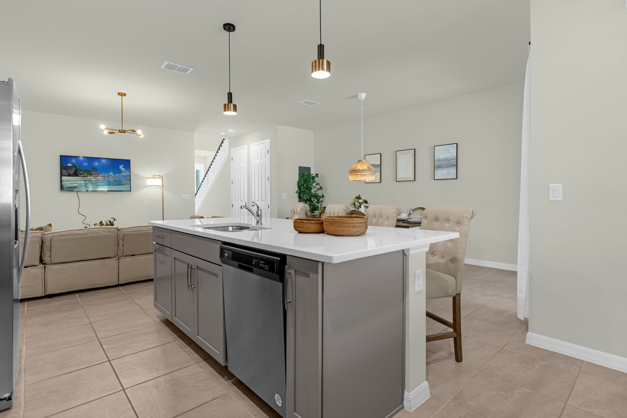 Open-concept kitchen island with a sink and dishwasher, featuring light gray cabinets and white countertops, overlooking a living area with a large TV and a dining area with a table and chairs.
