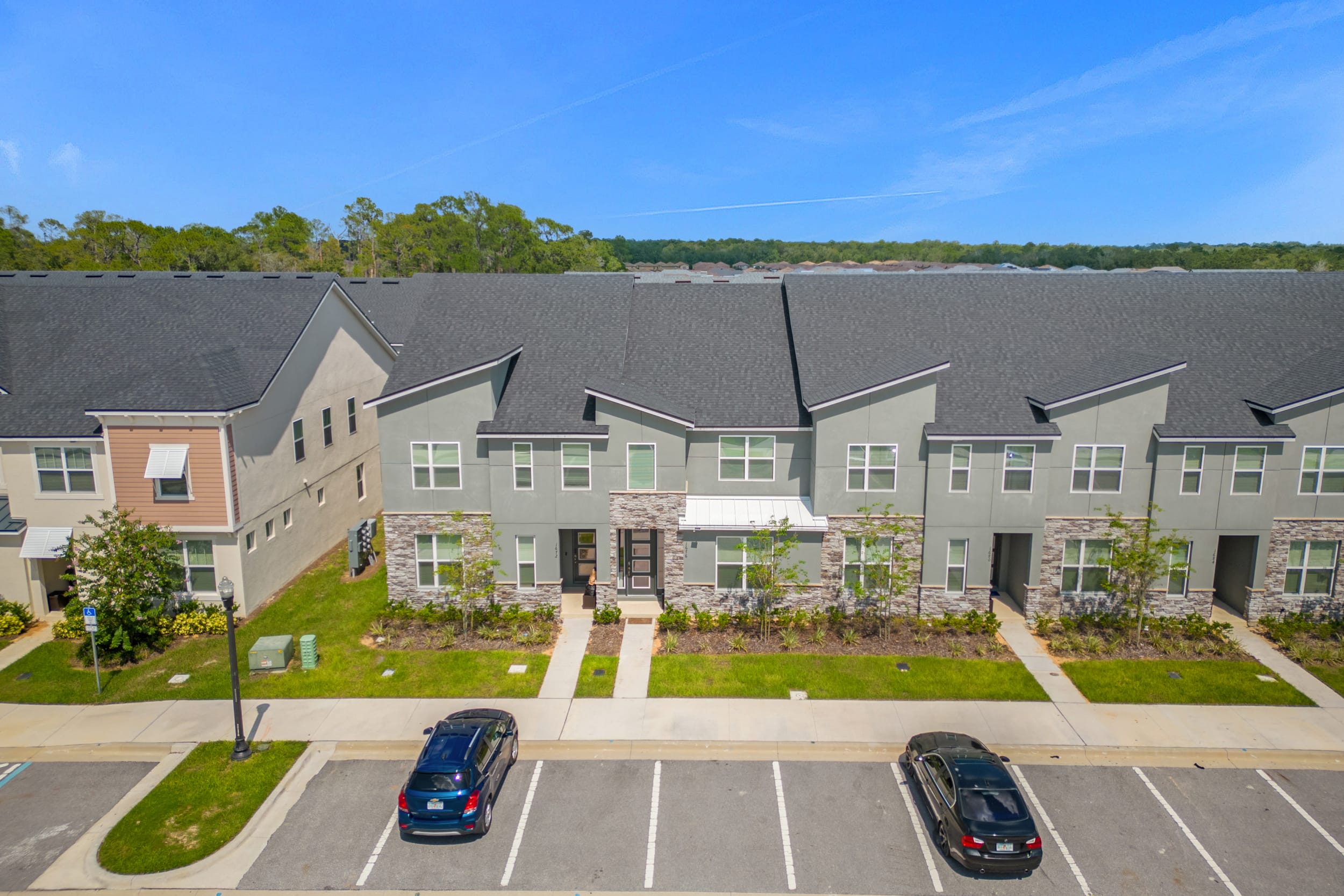 An aerial view showcasing a row of modern gray townhouses with stone accents and multiple windows, fronted by a paved parking lot with several cars. Green landscaping separates the buildings from the street.