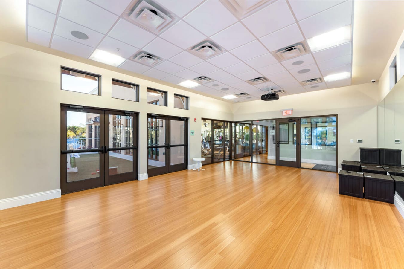 A spacious, brightly lit communal fitness or activity room with warm bamboo hardwood floors, a drop ceiling with recessed lighting, and large glass doors leading to an outdoor area and an adjacent gym. A projector is mounted on the ceiling.
