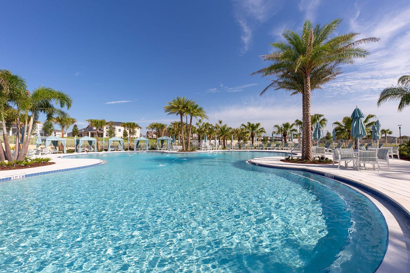 A large, curved communal swimming pool with clear blue water, surrounded by white decking, numerous palm trees, and light blue cabanas under a bright blue sky.