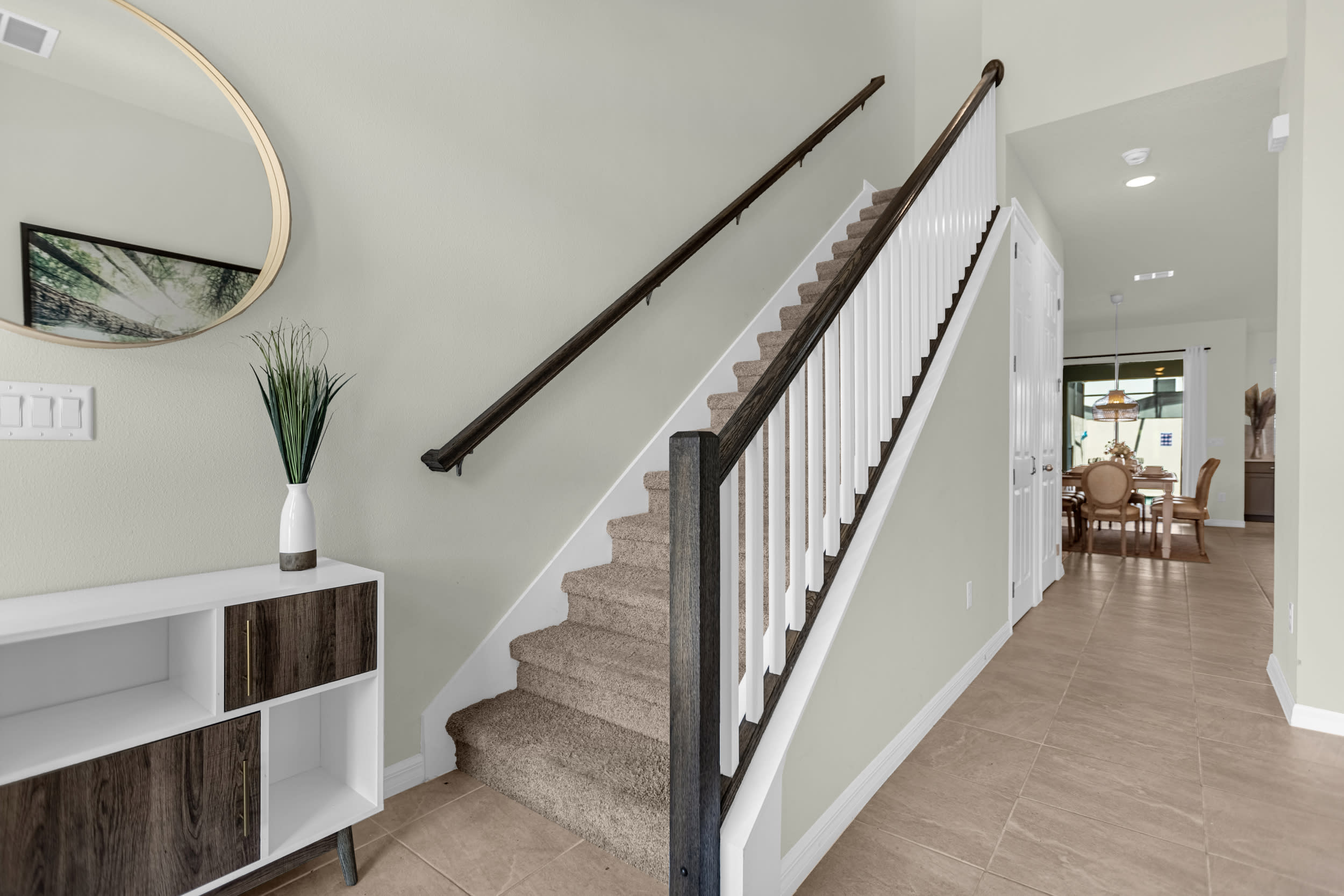 Entryway with light beige walls and tiled flooring, featuring a carpeted staircase with white balusters and dark wood handrails, and a decorative console table with a round mirror.