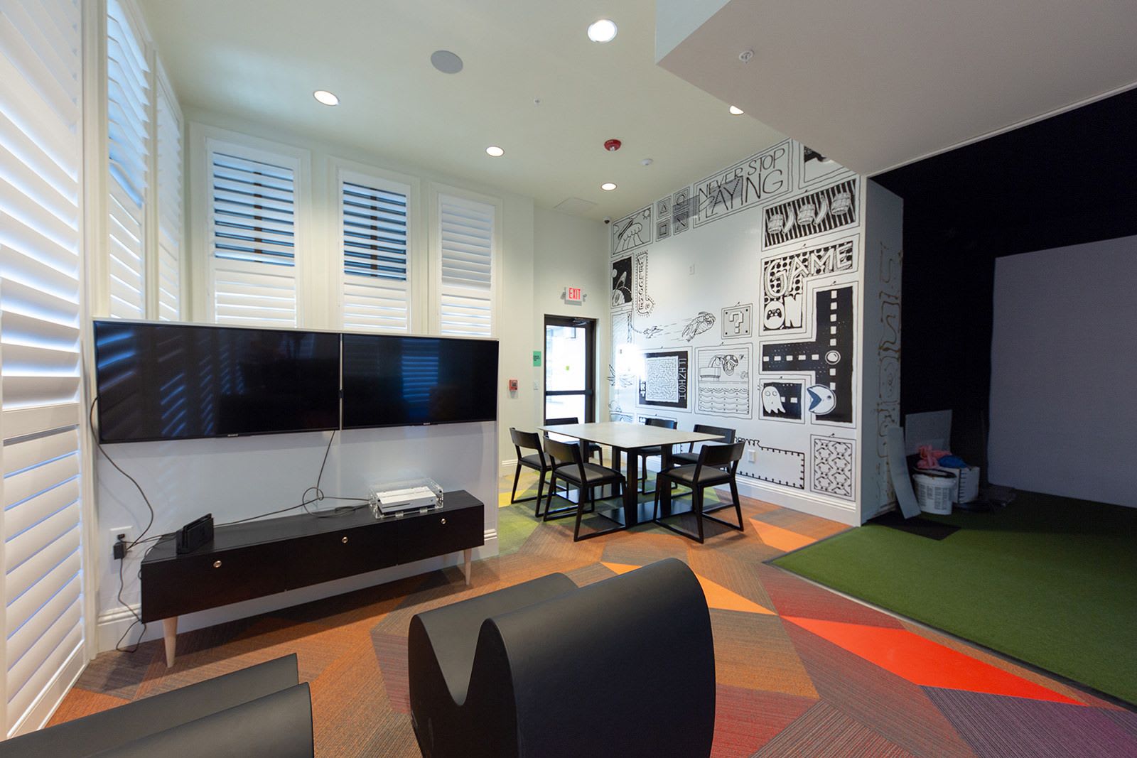 A contemporary indoor game room or lounge area featuring multiple large wall-mounted TVs, modern seating, a table with chairs, and a unique graphic wall mural, with a green turf area visible on the right.
