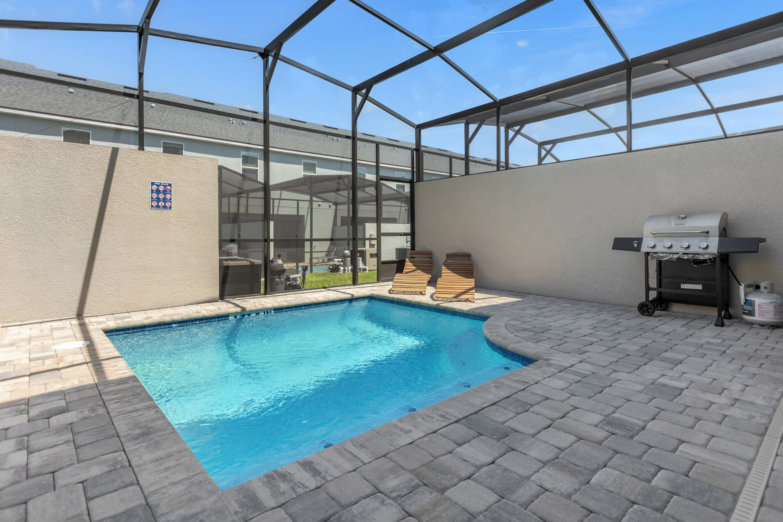 Screened-in private pool area with a rectangular pool, two wooden lounge chairs, and a BBQ grill. The patio features paver stones and is enclosed by a screen cage, offering an outdoor recreation space.