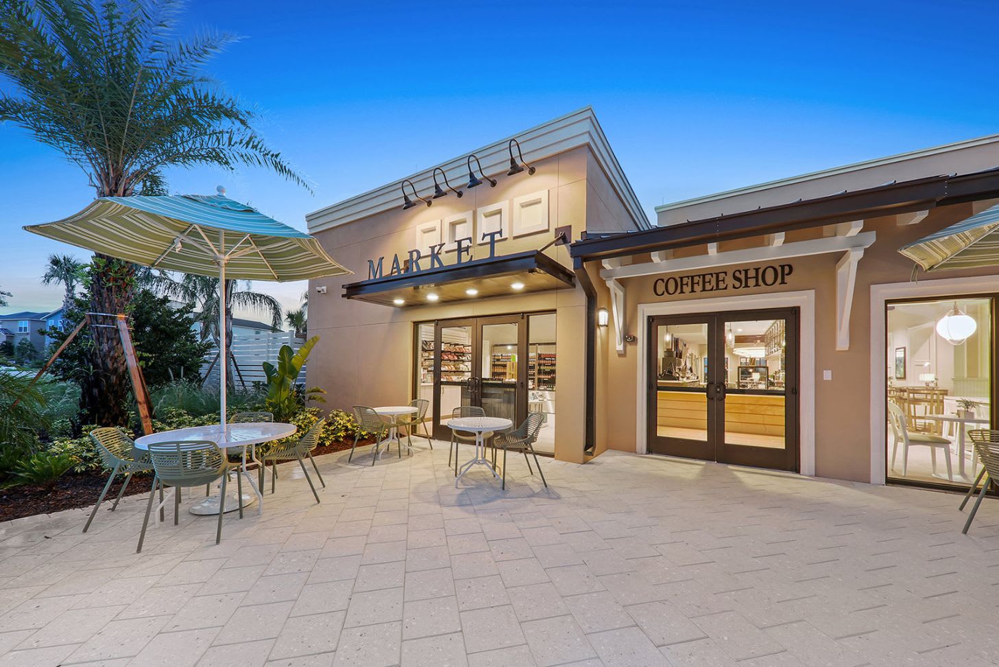 The modern exterior of a "MARKET" and "COFFEE SHOP" building with large glass doors, outdoor tables with umbrellas, and lush landscaping, suggesting communal amenities within the resort.