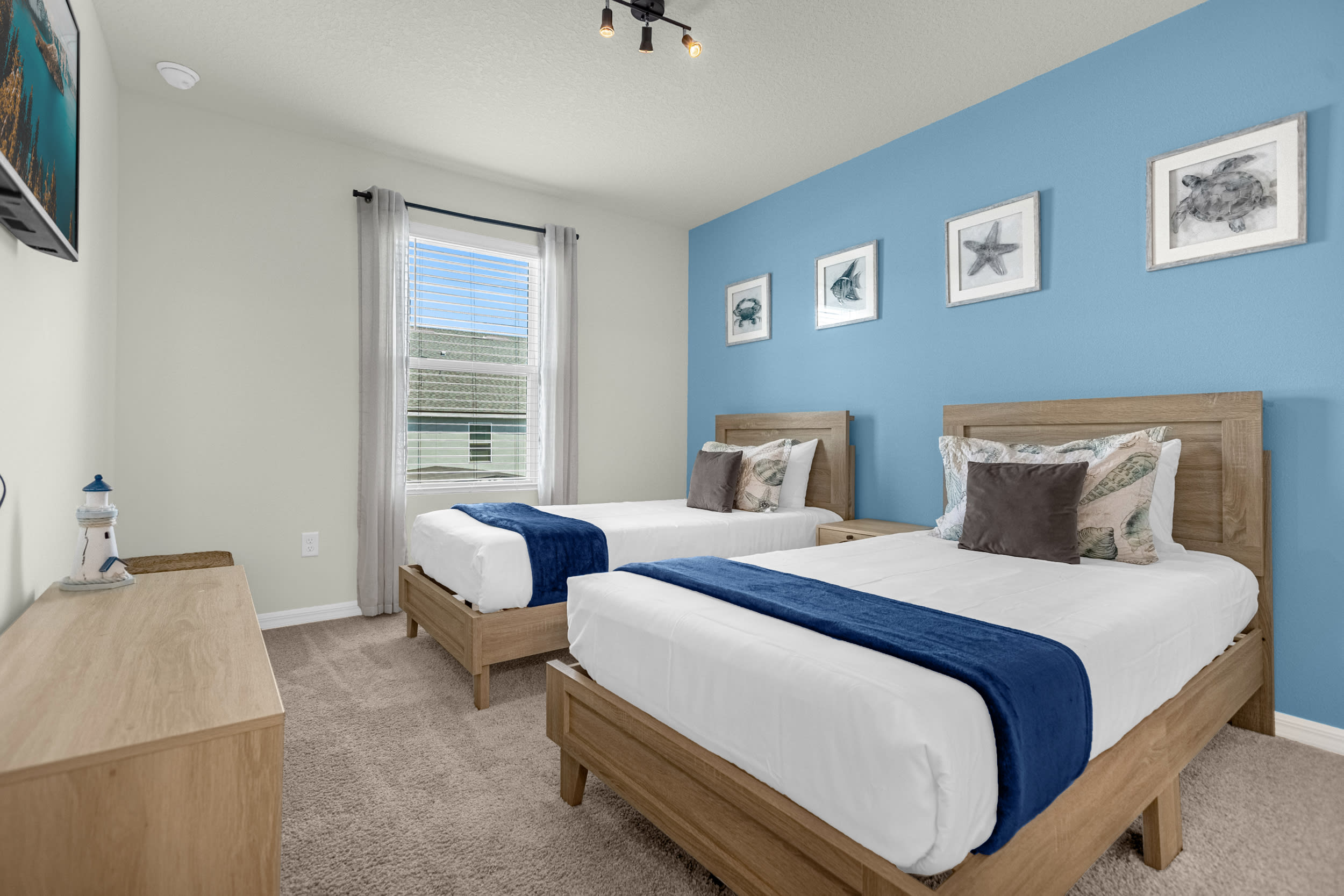 A bright bedroom with two twin beds featuring natural wood frames and blue accent throws, set against a vibrant blue accent wall. The room includes a window with blinds, a wooden dresser, and framed nautical-themed artwork.