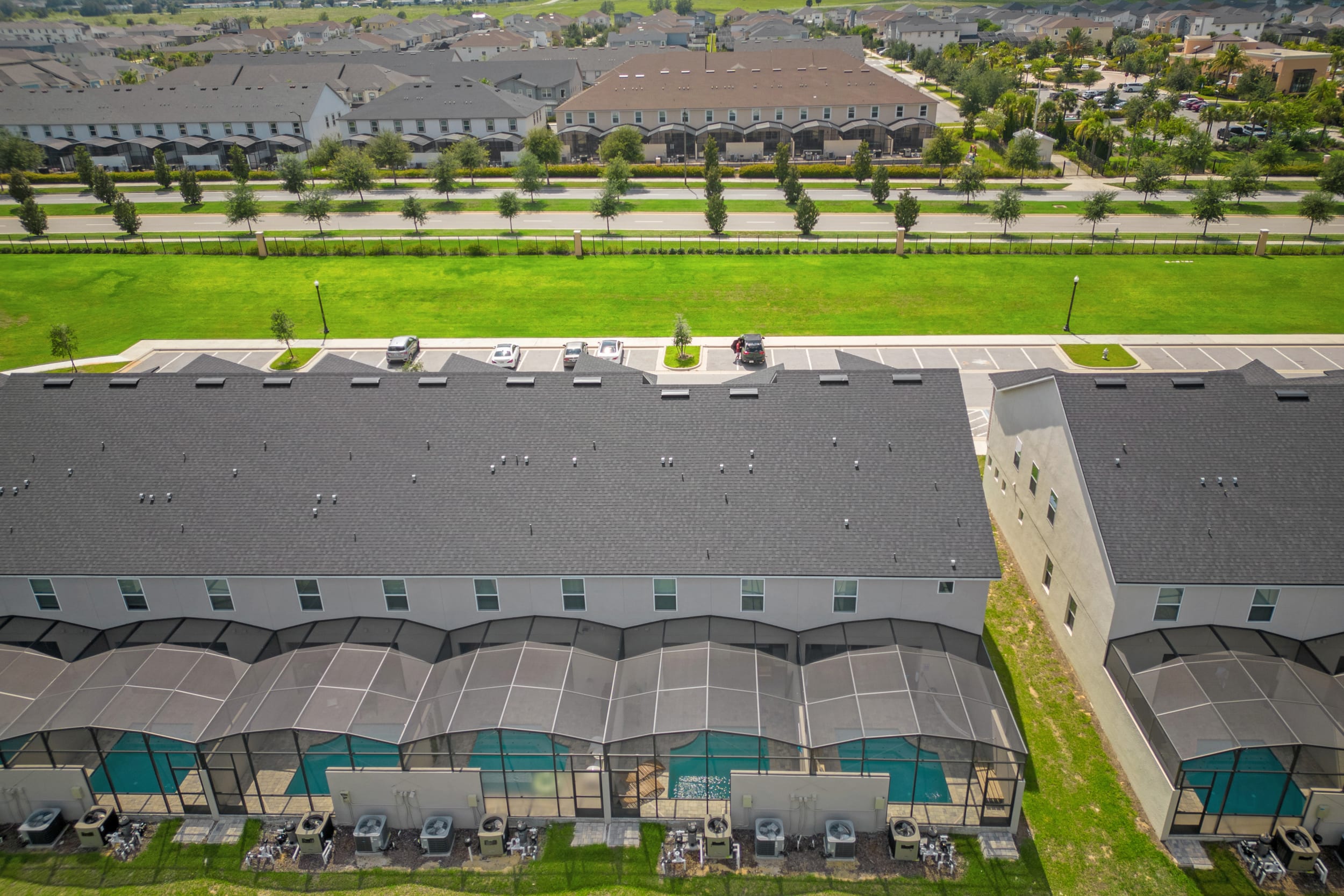 An aerial view of the rear of several townhouses, each featuring a private screened-in pool area with turquoise water. The properties are backed by a long green lawn and distant residential areas.