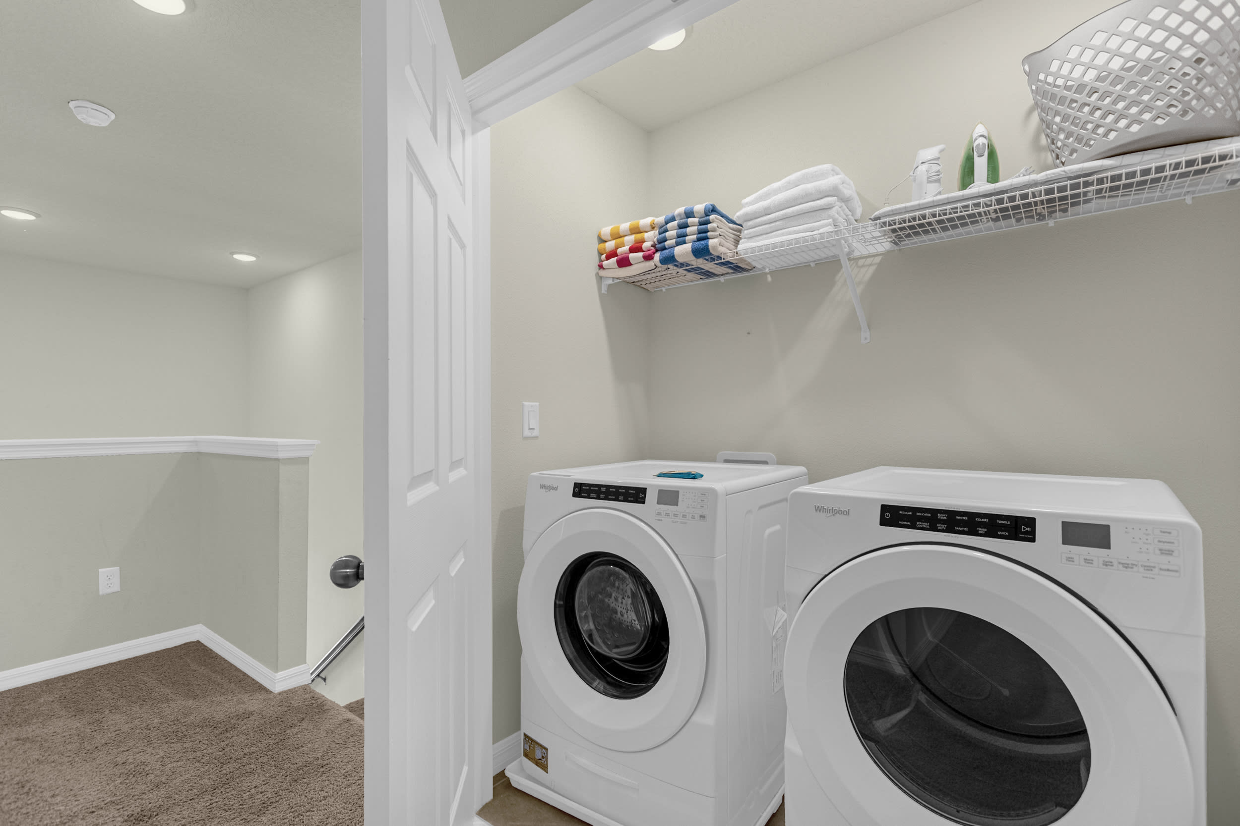 A dedicated laundry closet featuring a white front-loading washer and dryer set, with a wire shelf above holding neatly folded towels and laundry supplies. The space is well-lit and functional.