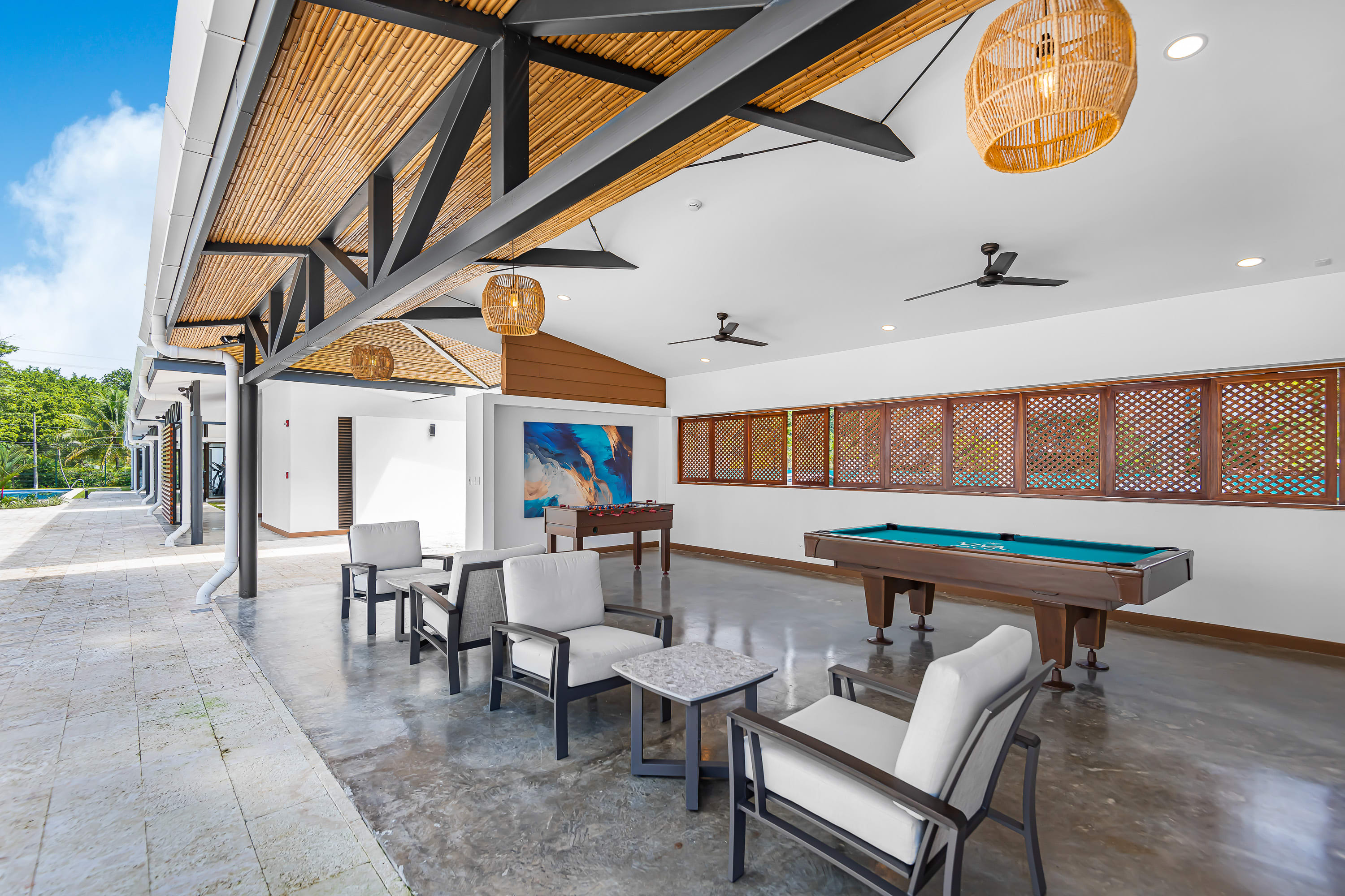Spacious covered outdoor game room featuring a pool table, foosball table, and several seating areas with armchairs and coffee tables, all under a high ceiling with bamboo accents and ceiling fans.