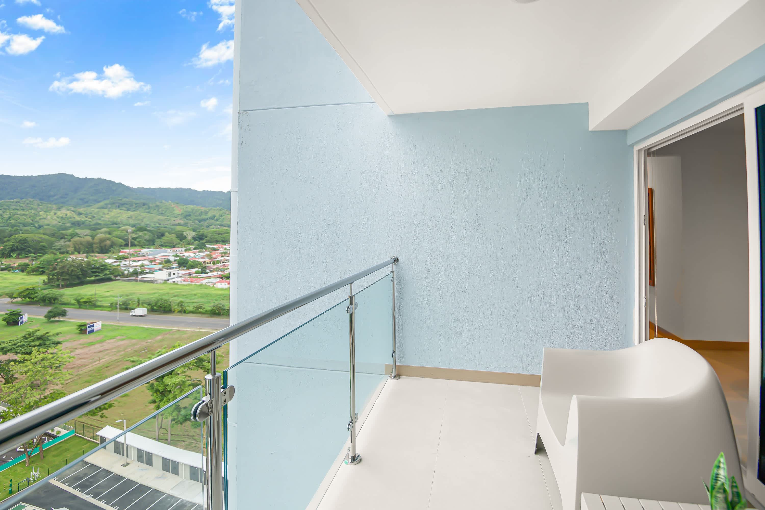 Balcony with a glass railing overlooking a vibrant green landscape with distant mountains and a town. A single white outdoor chair is visible, suggesting a quiet spot for relaxation.