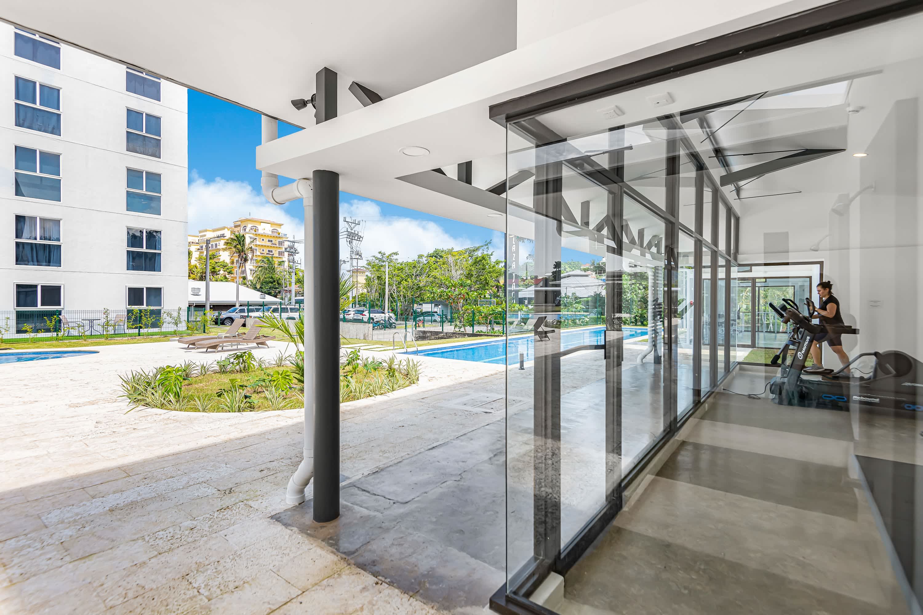 Modern outdoor communal area featuring a rectangular swimming pool surrounded by lush landscaping and paved walkways. Adjacent is a glass-enclosed gym with exercise equipment, where a person is visible on a treadmill.