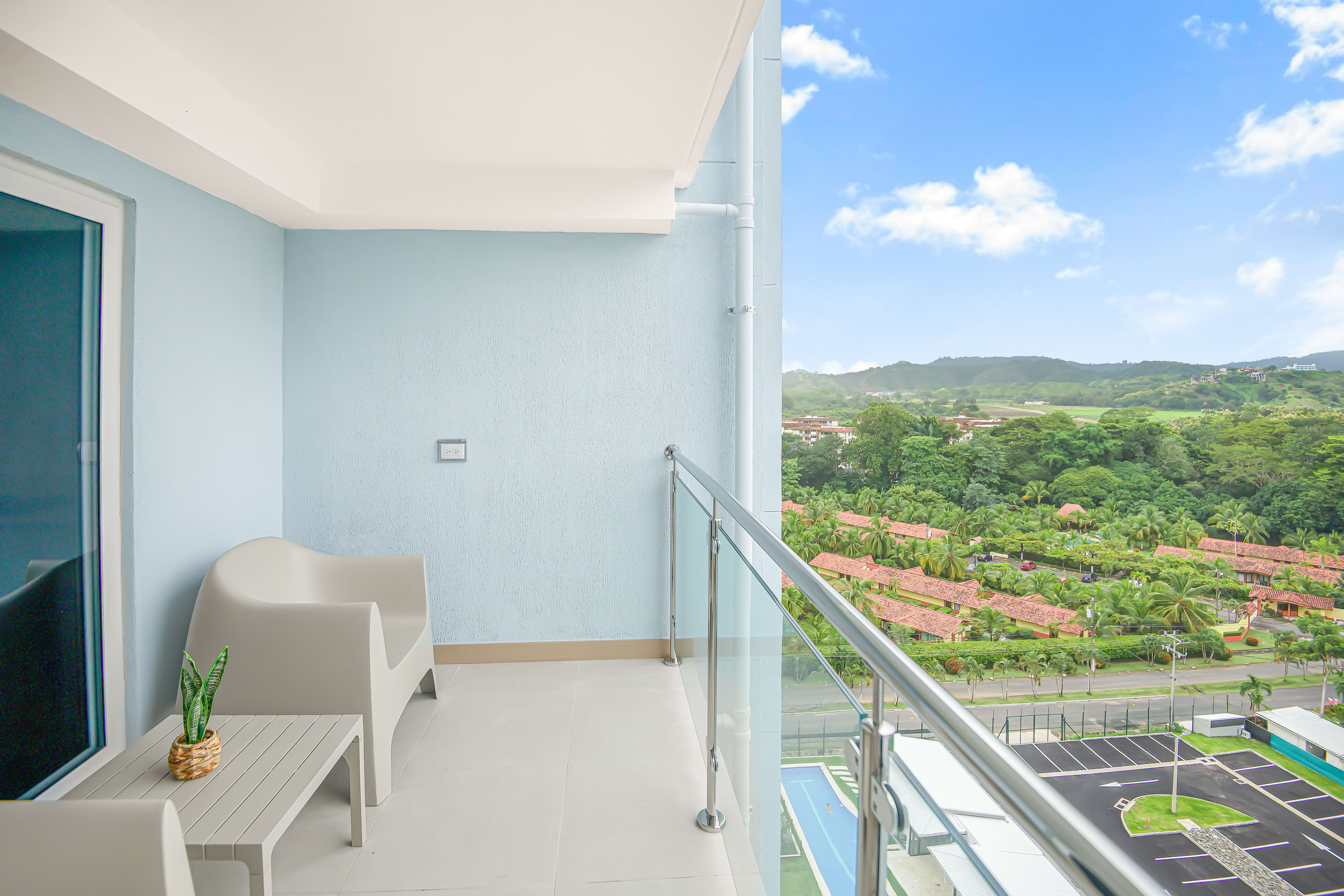 Spacious balcony with light blue textured walls and a glass railing, offering expansive views of lush green mountains and a residential area below. Two modern white outdoor chairs and a small table provide seating.