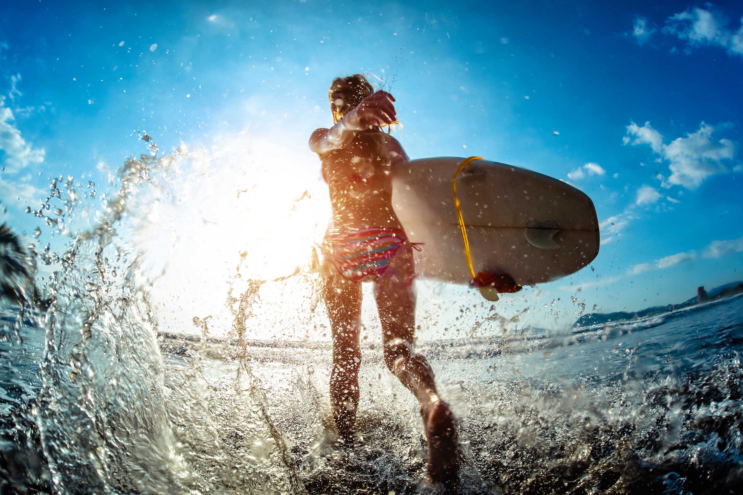 A person in a bikini, holding a surfboard, splashes through the shallow ocean water with the bright sun creating a strong backlight. Water sprays around them, highlighting the dynamic movement.