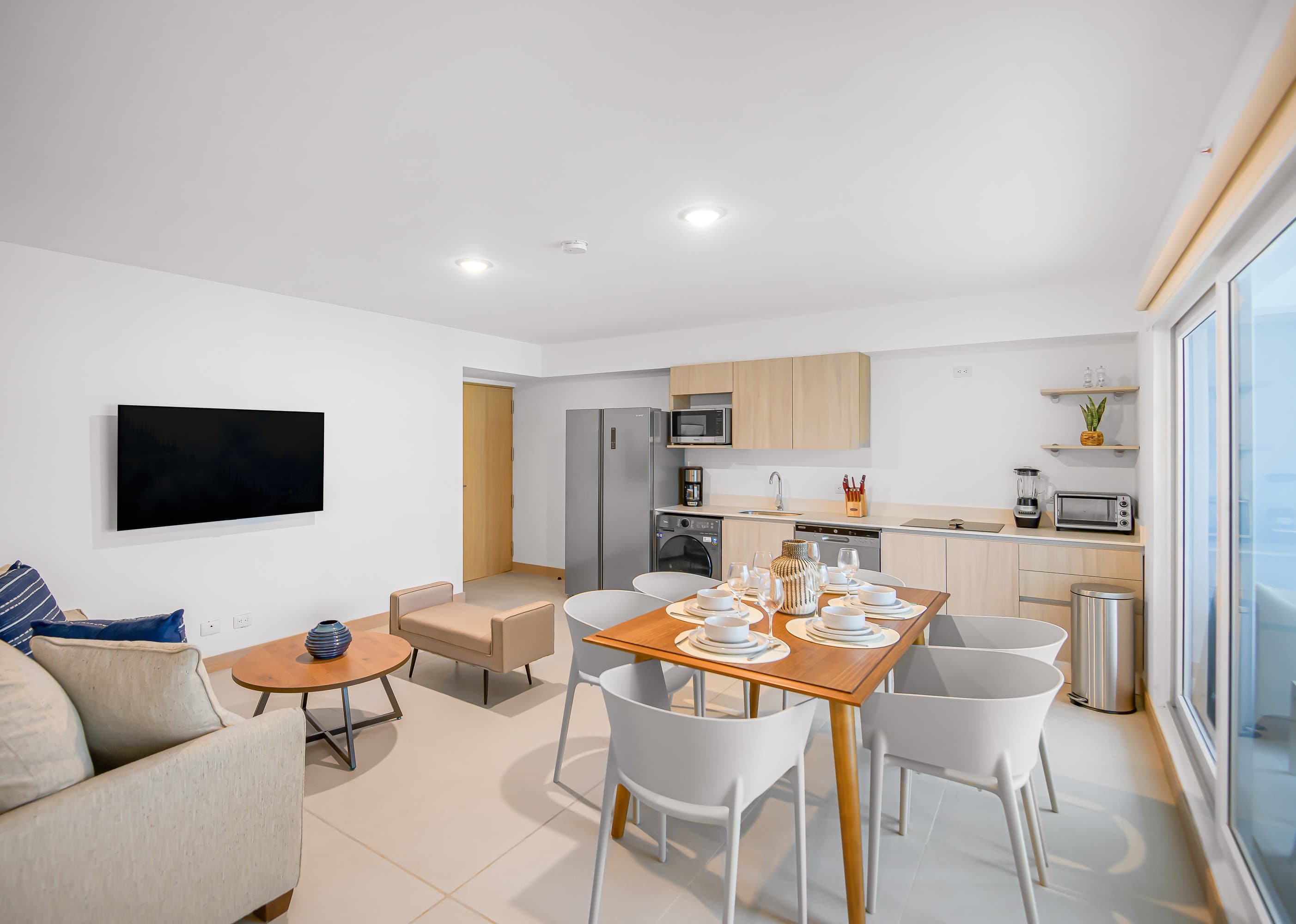 Spacious open-concept living room, dining area, and kitchen, featuring a light-colored sofa, a round coffee table, a wooden dining table, and a modern kitchen with stainless steel appliances and light wood cabinets.