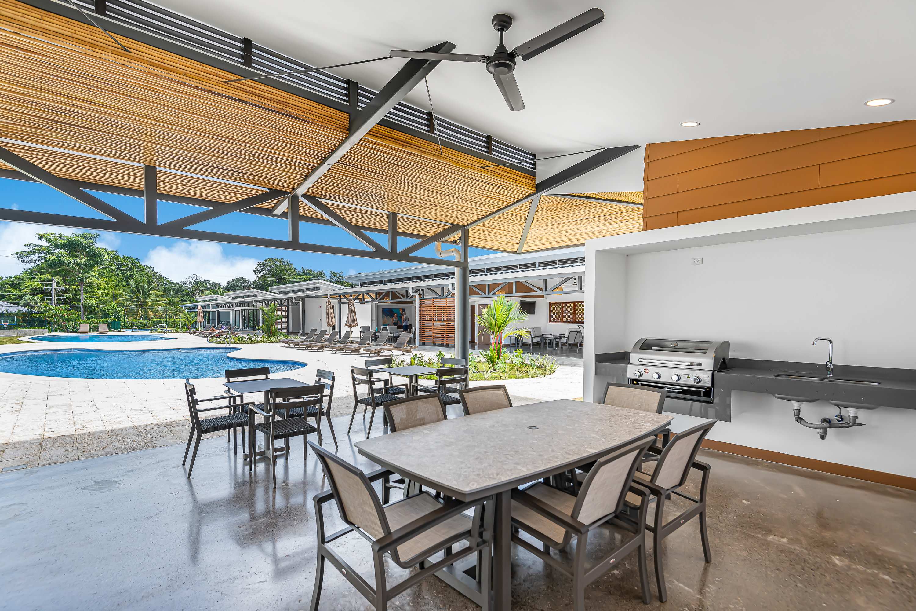 Covered outdoor dining and BBQ area with multiple tables and chairs, a built-in grill station with a sink, and a view of the swimming pool and lounge area in the background.