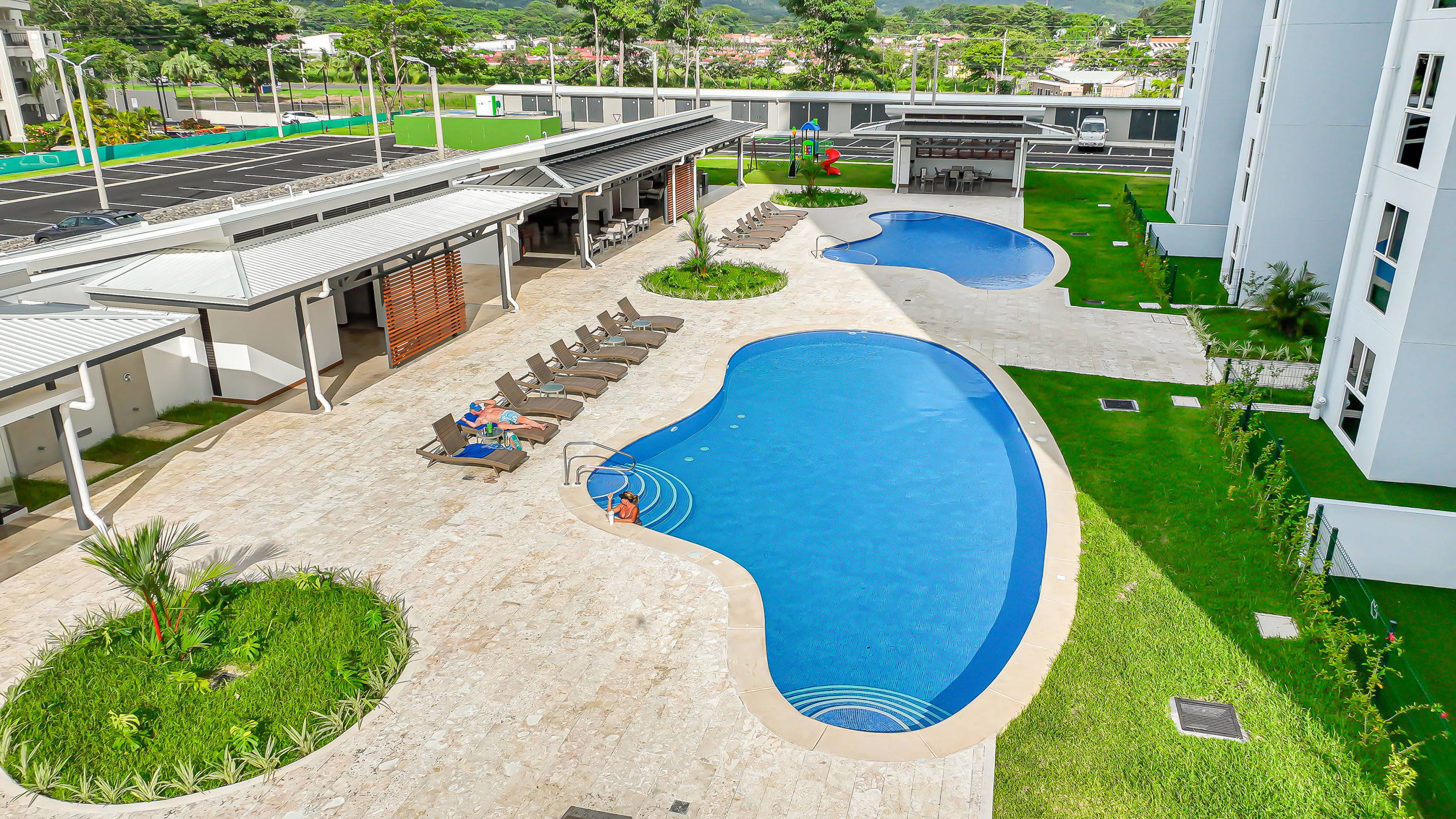 Elevated view of a resort-style outdoor area featuring two irregularly shaped swimming pools surrounded by light-colored paving, lounge chairs, and manicured green lawns, with residential buildings in the background.