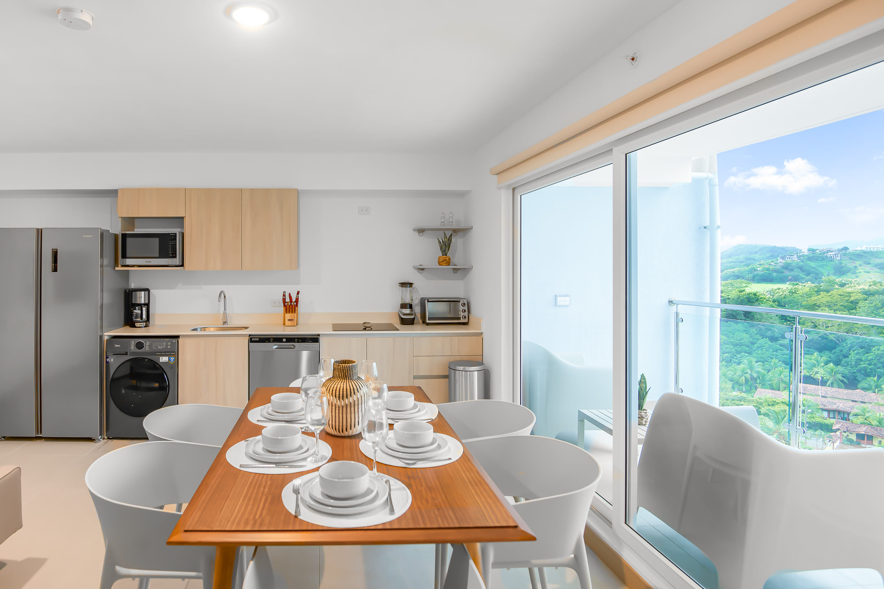 Open-concept dining area with a rectangular wooden table set for six, surrounded by modern white chairs, adjacent to a contemporary kitchen. Large sliding glass doors lead to a balcony with mountain views.
