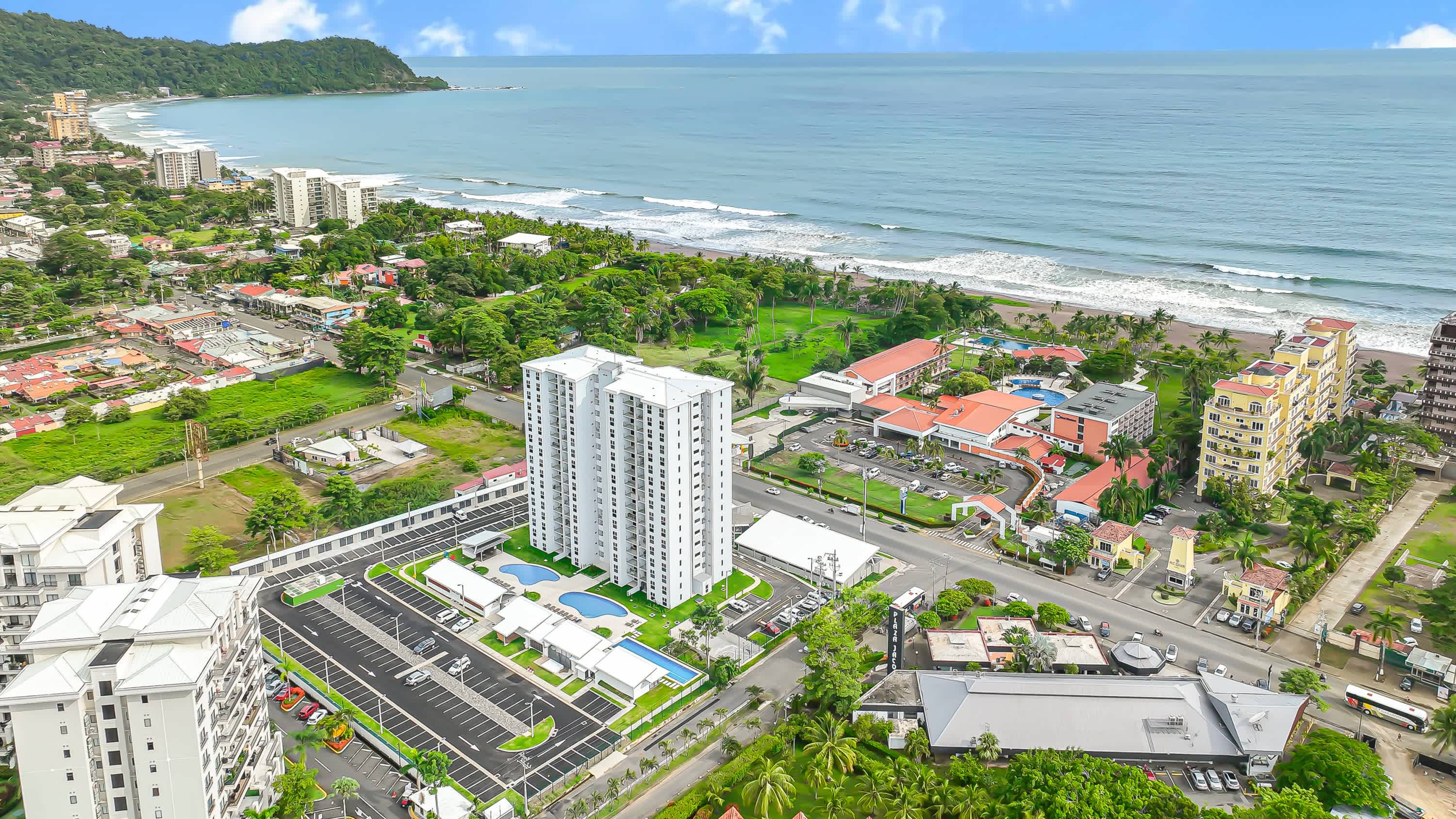High aerial view showcasing a modern white high-rise condominium complex with multiple swimming pools and parking lots, situated along a sandy beach with ocean waves and lush green landscape.