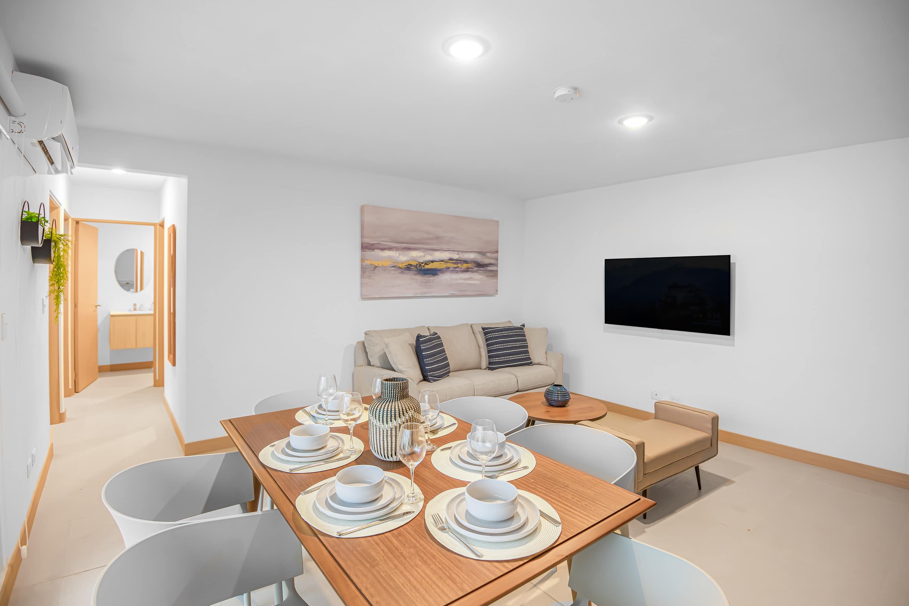 Contemporary dining area with a wooden table set for six, adjacent to a living room with a light-colored sofa and a wall-mounted television, leading to a hallway with a bathroom visible.