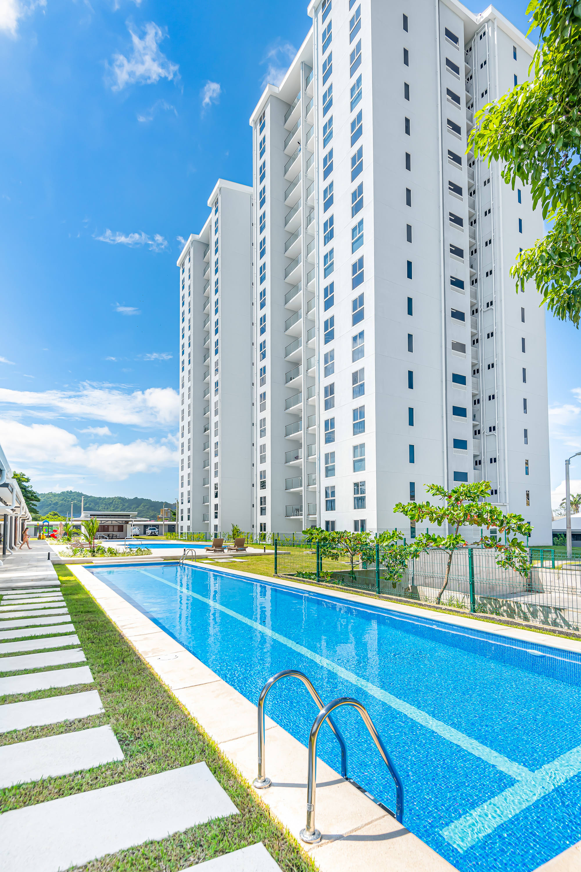 Ground-level view of a long, rectangular swimming pool with blue tiles and metal steps, surrounded by a green lawn and white stepping stones, with modern white high-rise buildings under a clear blue sky.