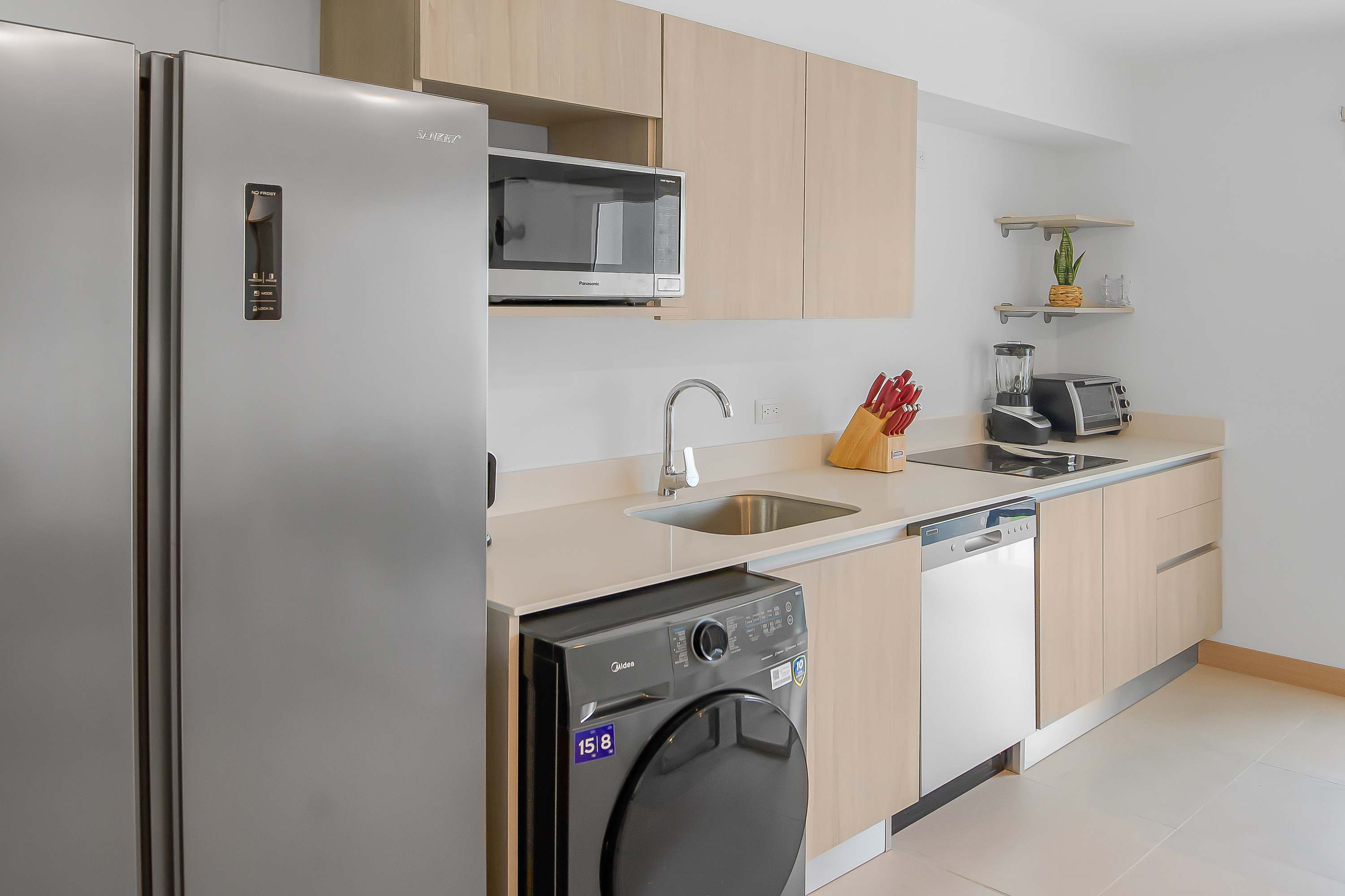 Modern kitchen with light wood cabinetry, light countertops, and stainless steel appliances including a large refrigerator, microwave, dishwasher, and a built-in washer/dryer unit. A sink with a gooseneck faucet is centered under the microwave.