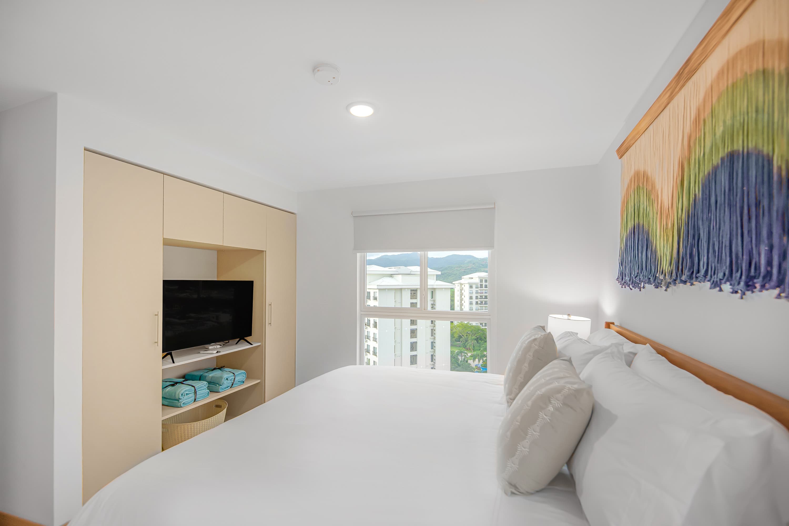 Bright bedroom featuring a large bed with white bedding and light blue pillows, built-in light wood cabinetry with a TV niche, and a window offering city and mountain views. A colorful macrame wall hanging adds a decorative touch.