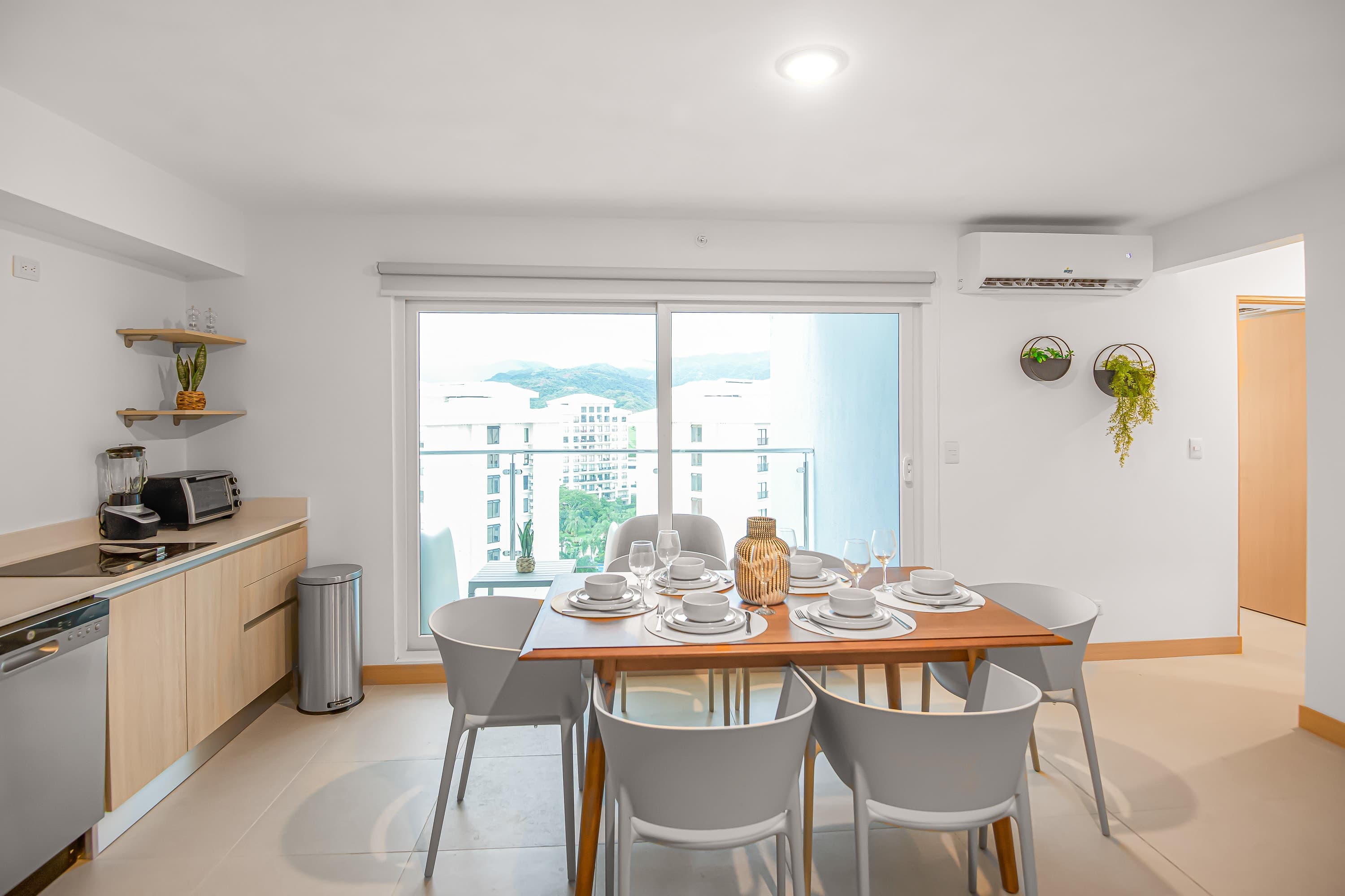 Bright kitchen and dining space with light wood cabinetry, stainless steel appliances, a wooden dining table set for six, and a large sliding glass door offering views of the exterior and mountains.