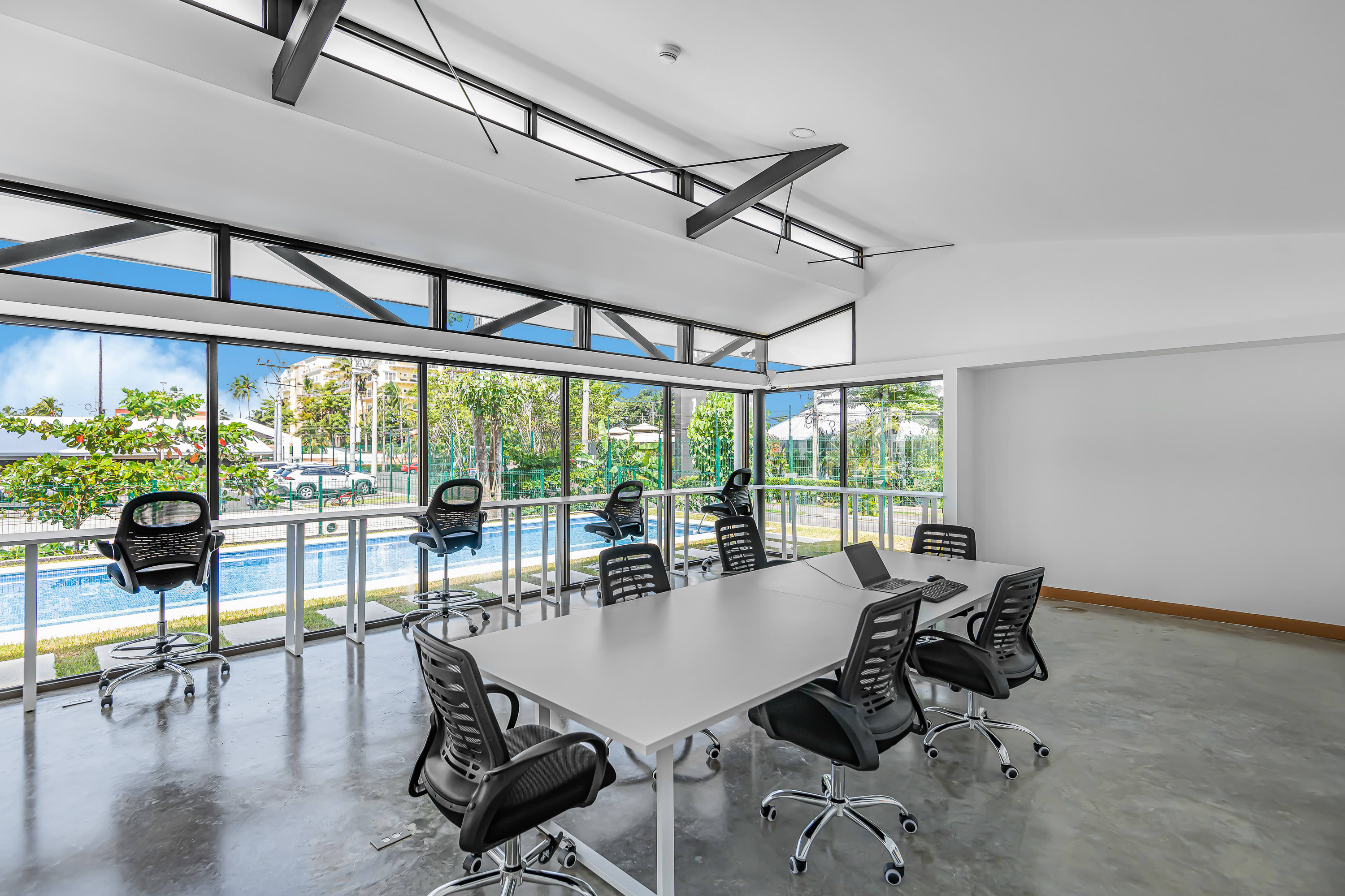 Bright, modern co-working space with a long white conference table and multiple black office chairs. Large windows offer a panoramic view of an outdoor pool area and tropical trees under a blue sky.