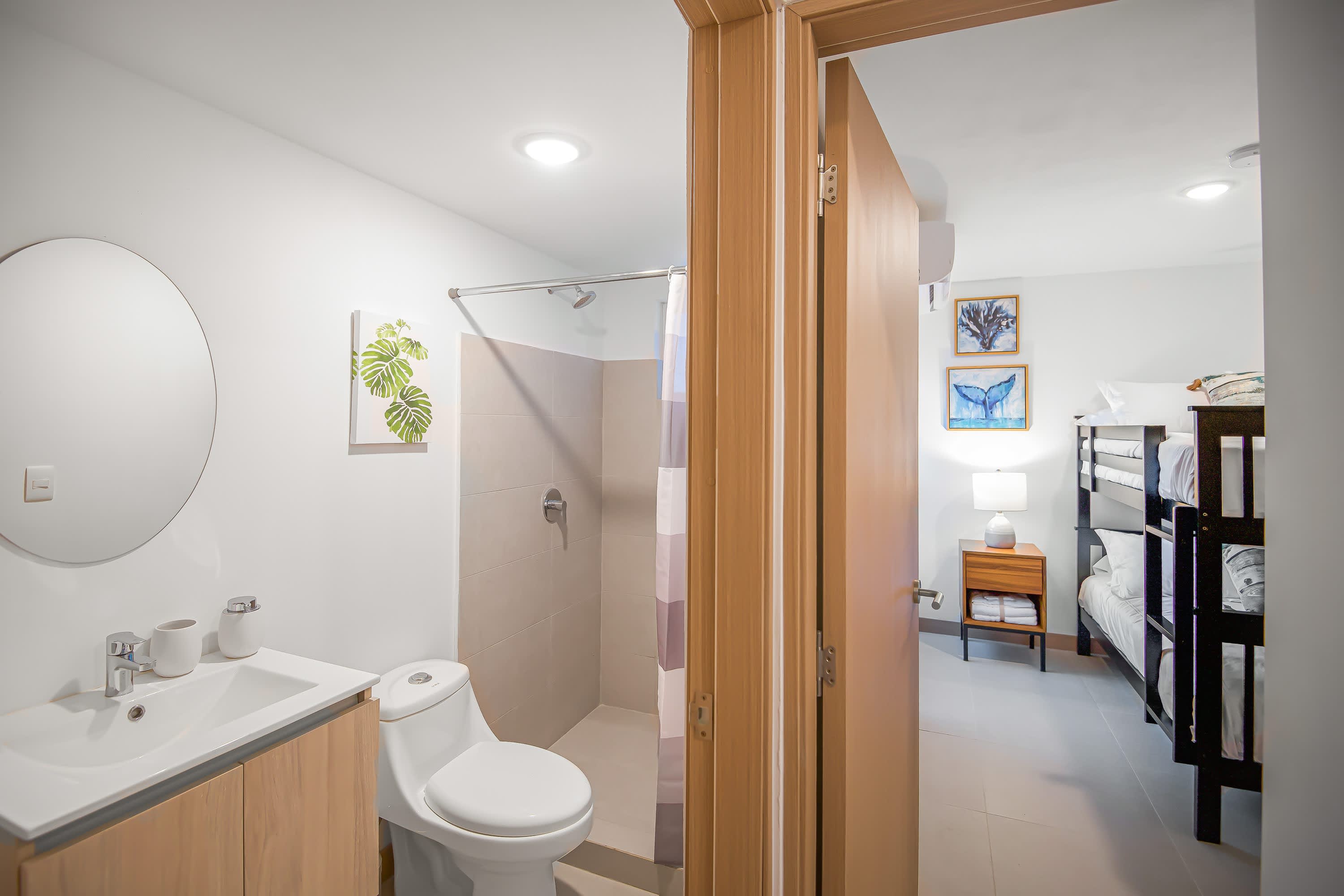 Modern bathroom featuring a white vanity with a rectangular sink, a round mirror, and a toilet, with a tiled shower stall visible. The adjacent room has bunk beds.