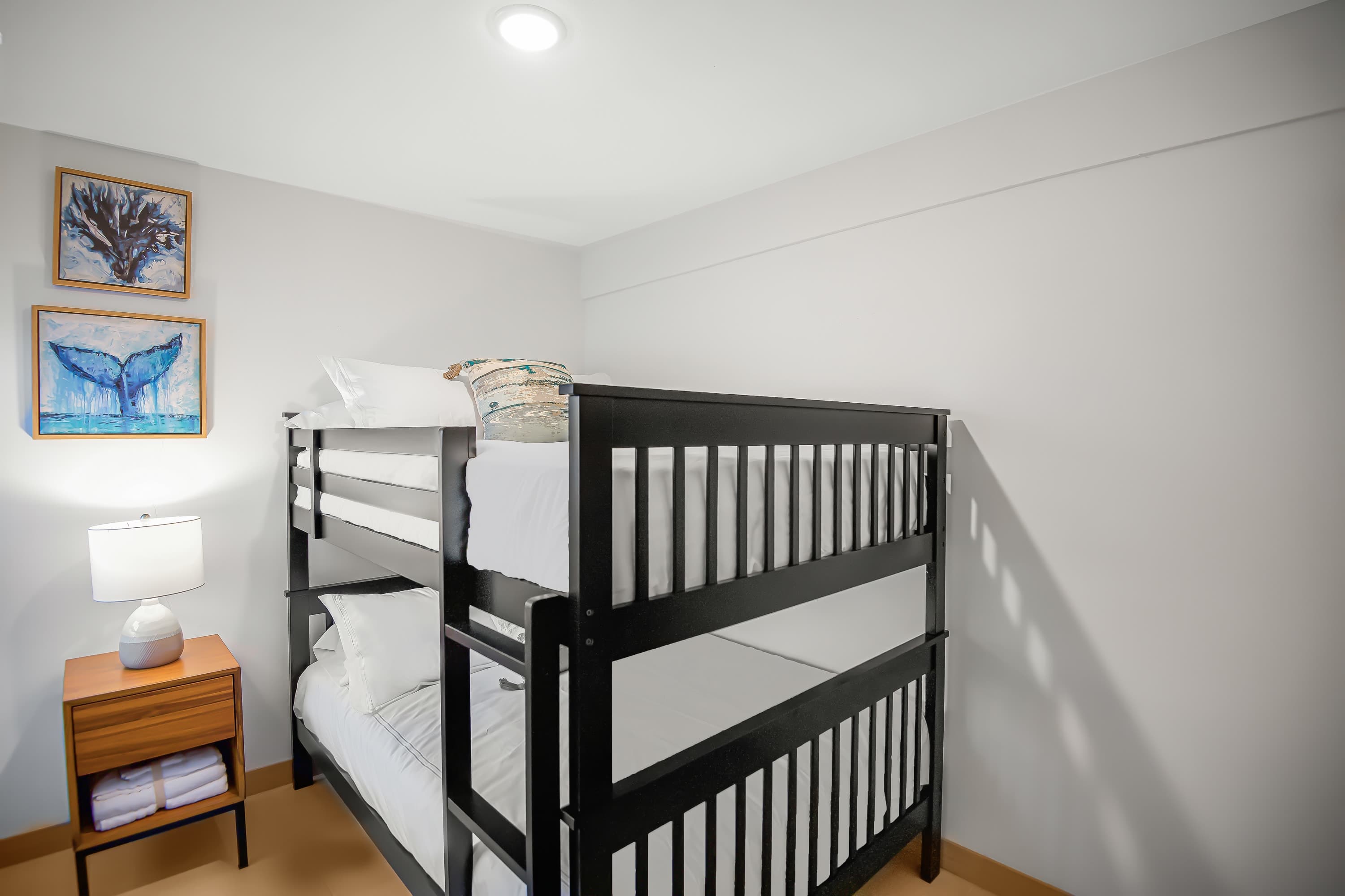 Bright bedroom with white walls featuring a black wooden bunk bed with white bedding, a wooden nightstand with a lamp, and two framed ocean-themed artworks on the wall.