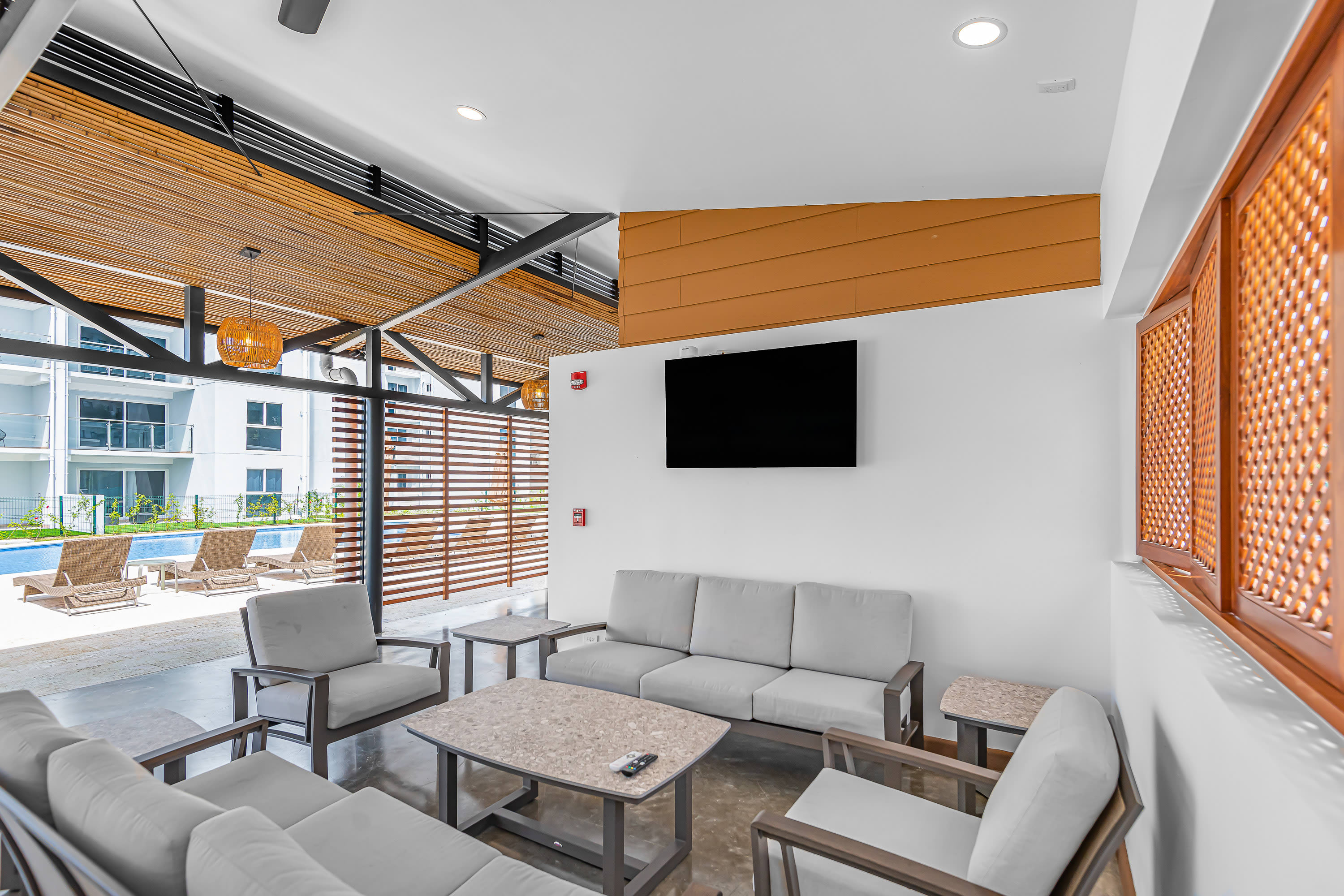 Covered outdoor lounge area with comfortable grey sofas and coffee tables, featuring a wall-mounted TV, slatted wooden screens, and a view of the swimming pool area in the background.