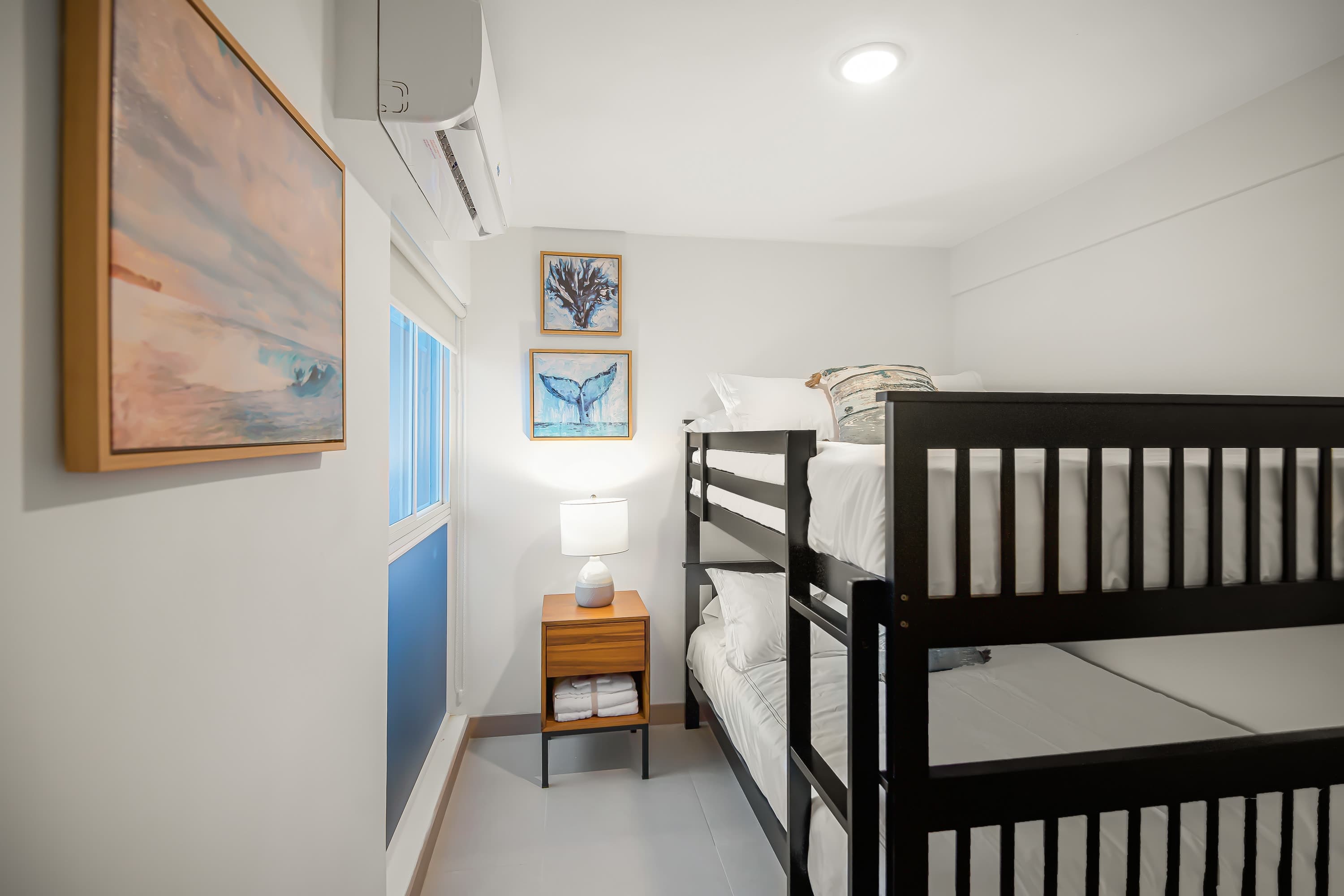 Cozy bedroom with white walls and a window, featuring a black wooden bunk bed, a wooden nightstand with a lamp, and ocean-themed artwork, with an air conditioning unit mounted above the window.