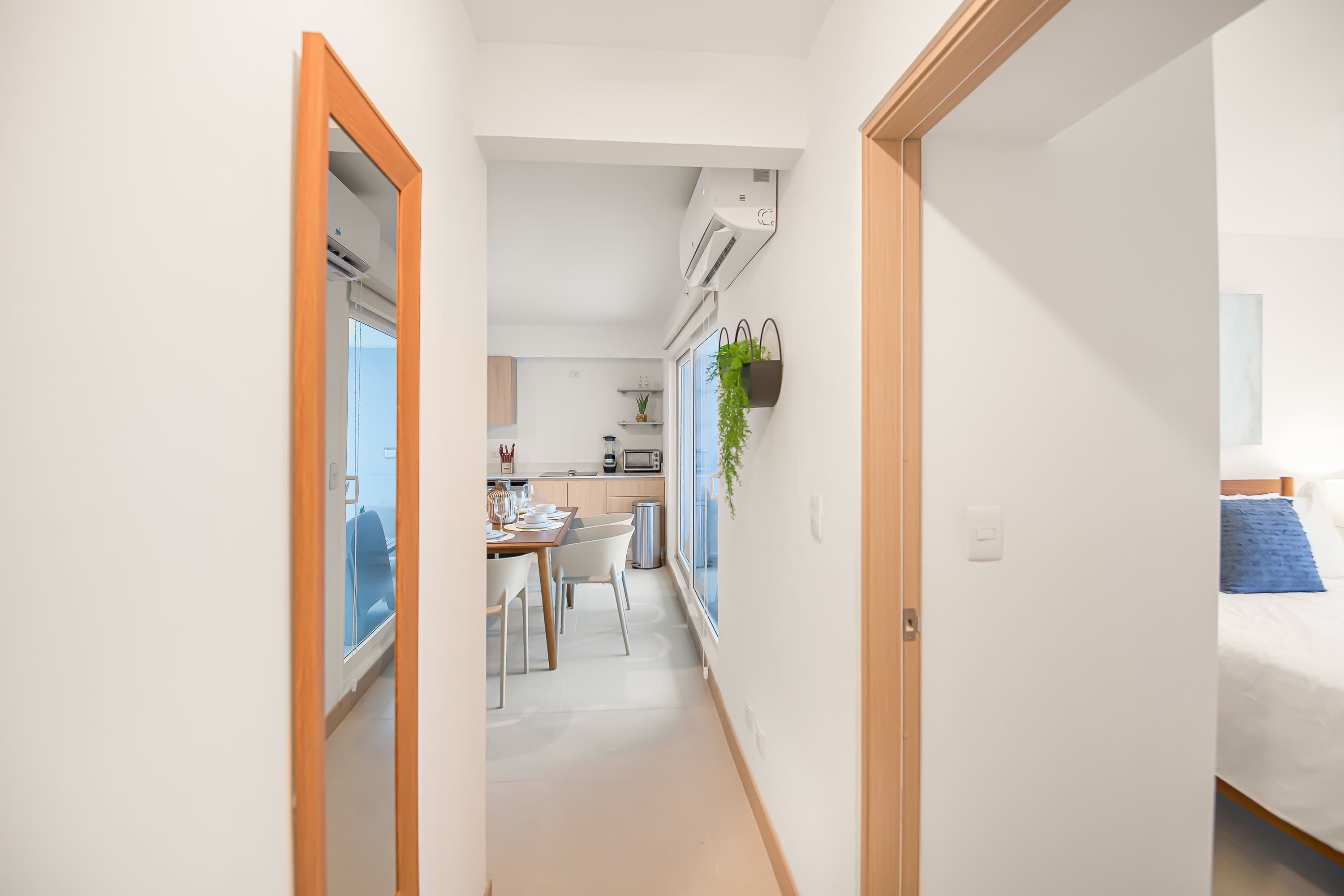 Bright hallway with white walls and light wood trim, featuring a full-length mirror on one side and leading towards the kitchen and dining area. An air conditioning unit is mounted high on the wall.