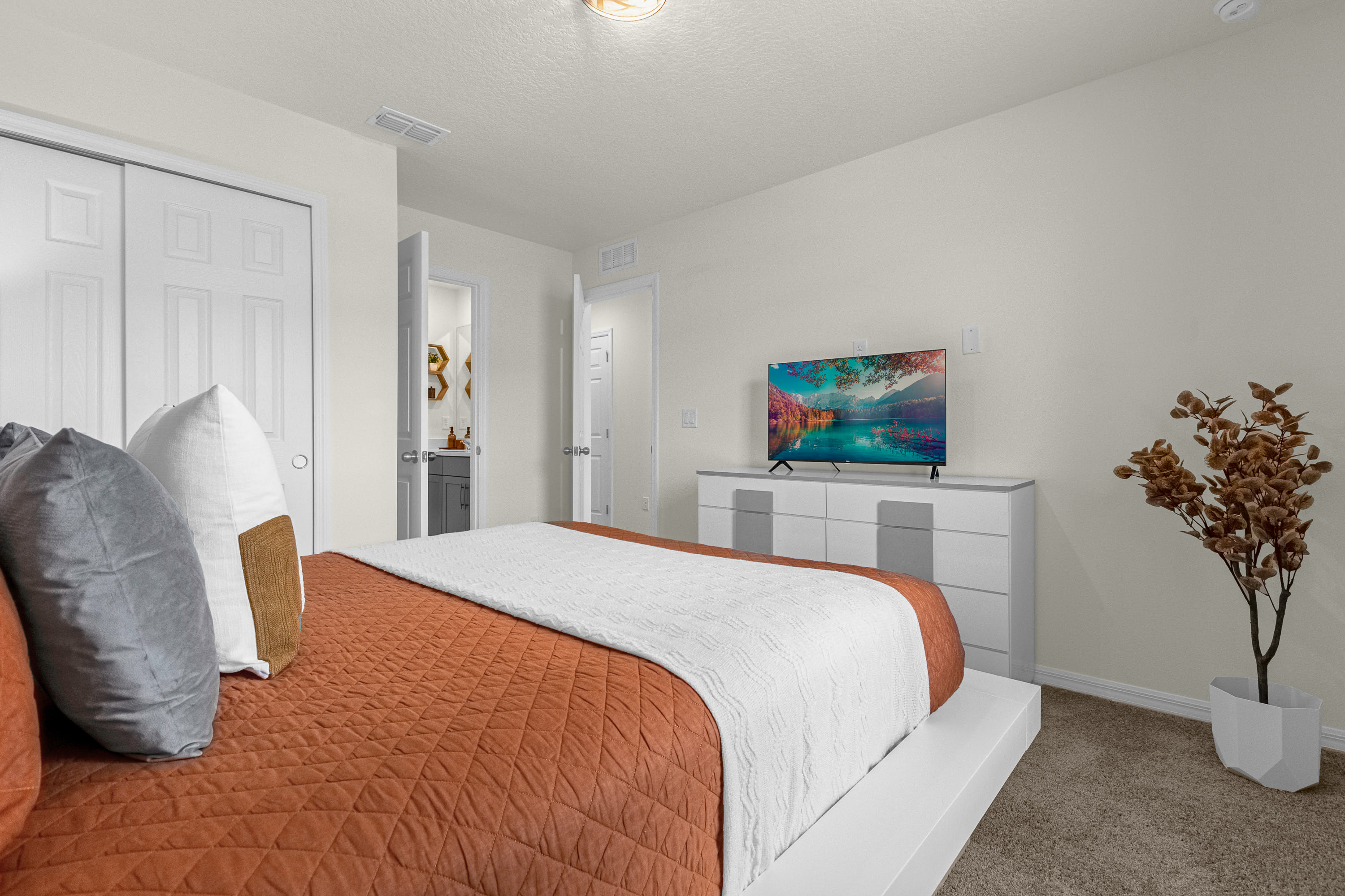 View of a modern bedroom with a white platform bed, rust and white bedding, a white dresser with a television, and a glimpse into an en-suite bathroom through an open door.