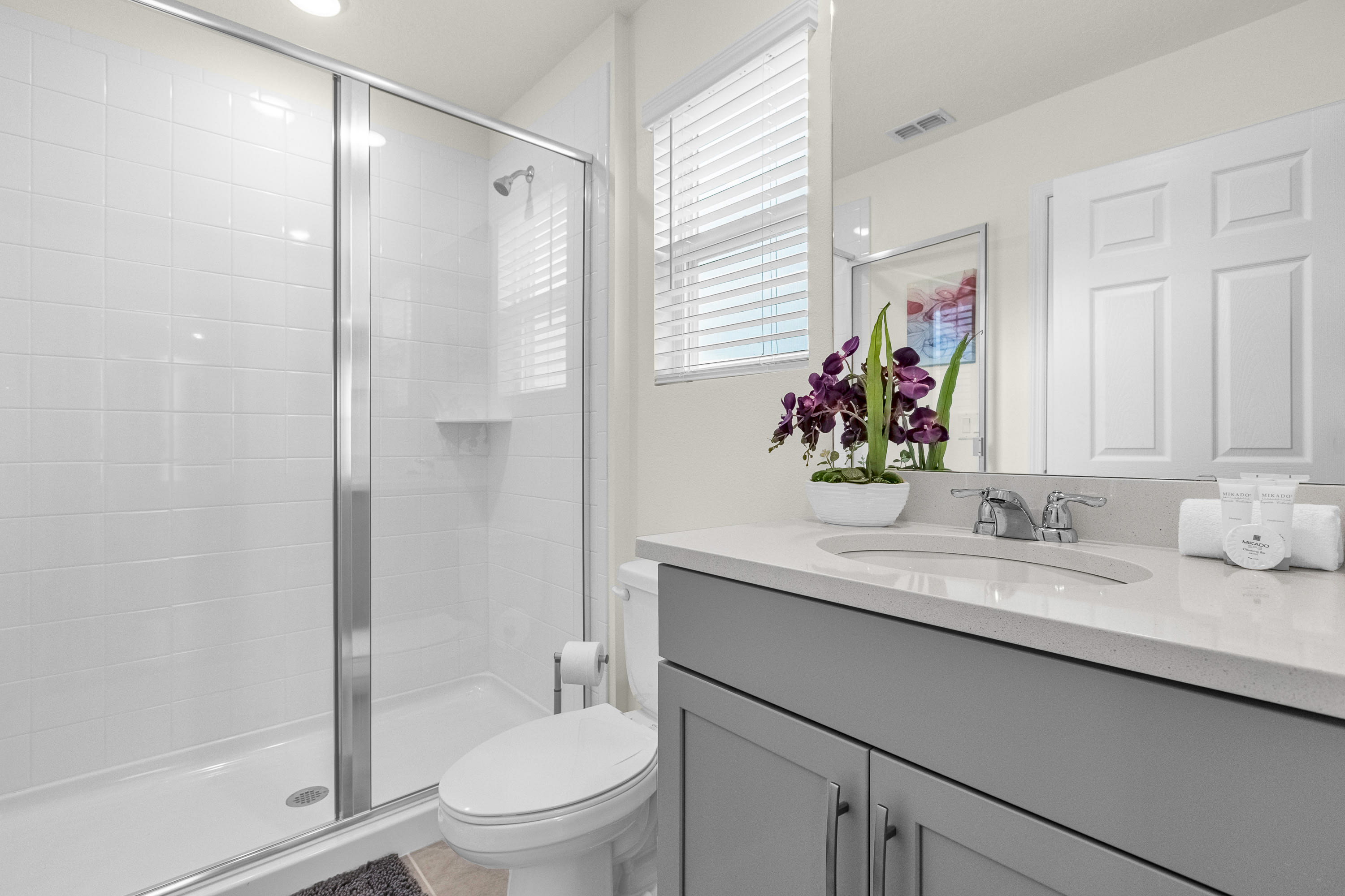 Modern bathroom featuring a white tiled walk-in shower with a glass enclosure, a gray vanity with a white quartz countertop and integrated sink, and a toilet. A window with white blinds provides natural light.