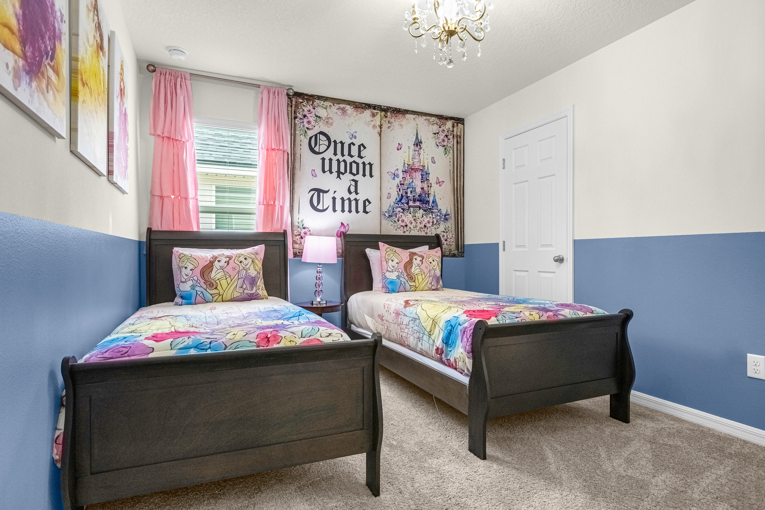 Charming kids' bedroom with two dark wood twin beds adorned with colorful Disney Princess bedding and pillows. A large 'Once Upon a Time' tapestry hangs above the beds, complementing the two-tone blue and white walls.