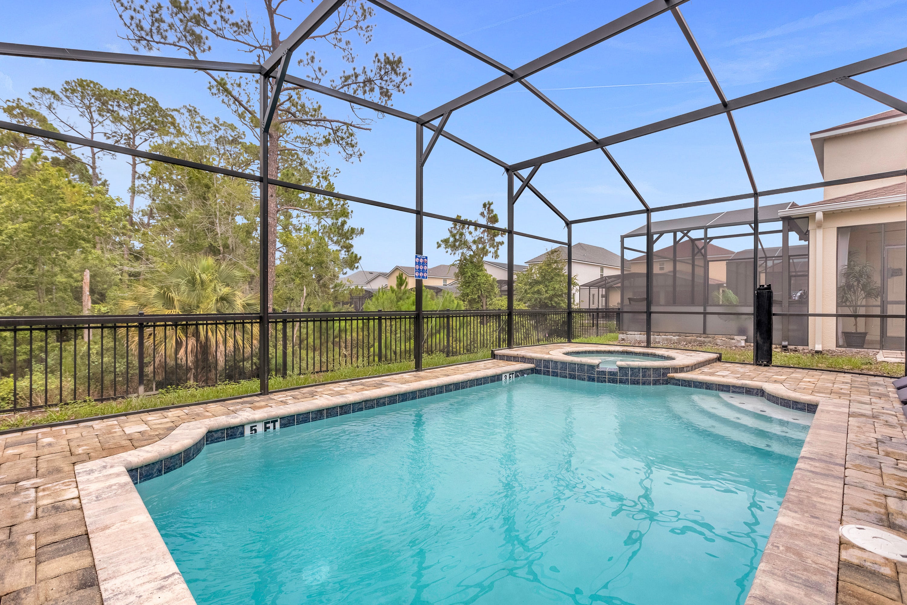 A screened-in outdoor pool area features a rectangular swimming pool with a 5-foot depth marker and an adjacent hot tub, surrounded by a paved deck and a black metal fence backing onto a wooded area.