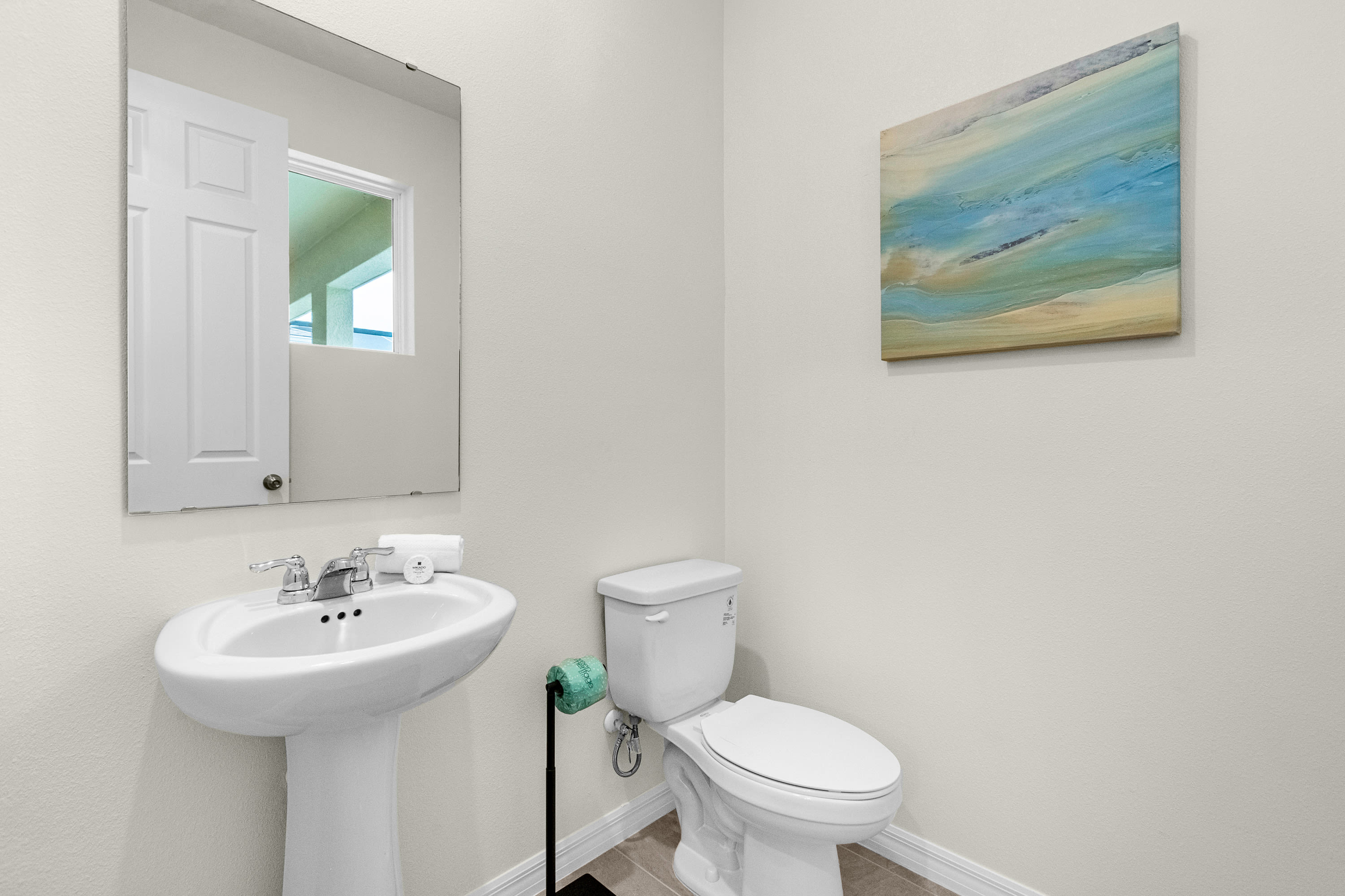 Compact powder room featuring a white pedestal sink with a chrome faucet and a toilet. A large rectangular mirror reflects a doorway and window, while an abstract painting adds a pop of color to the light-colored walls.