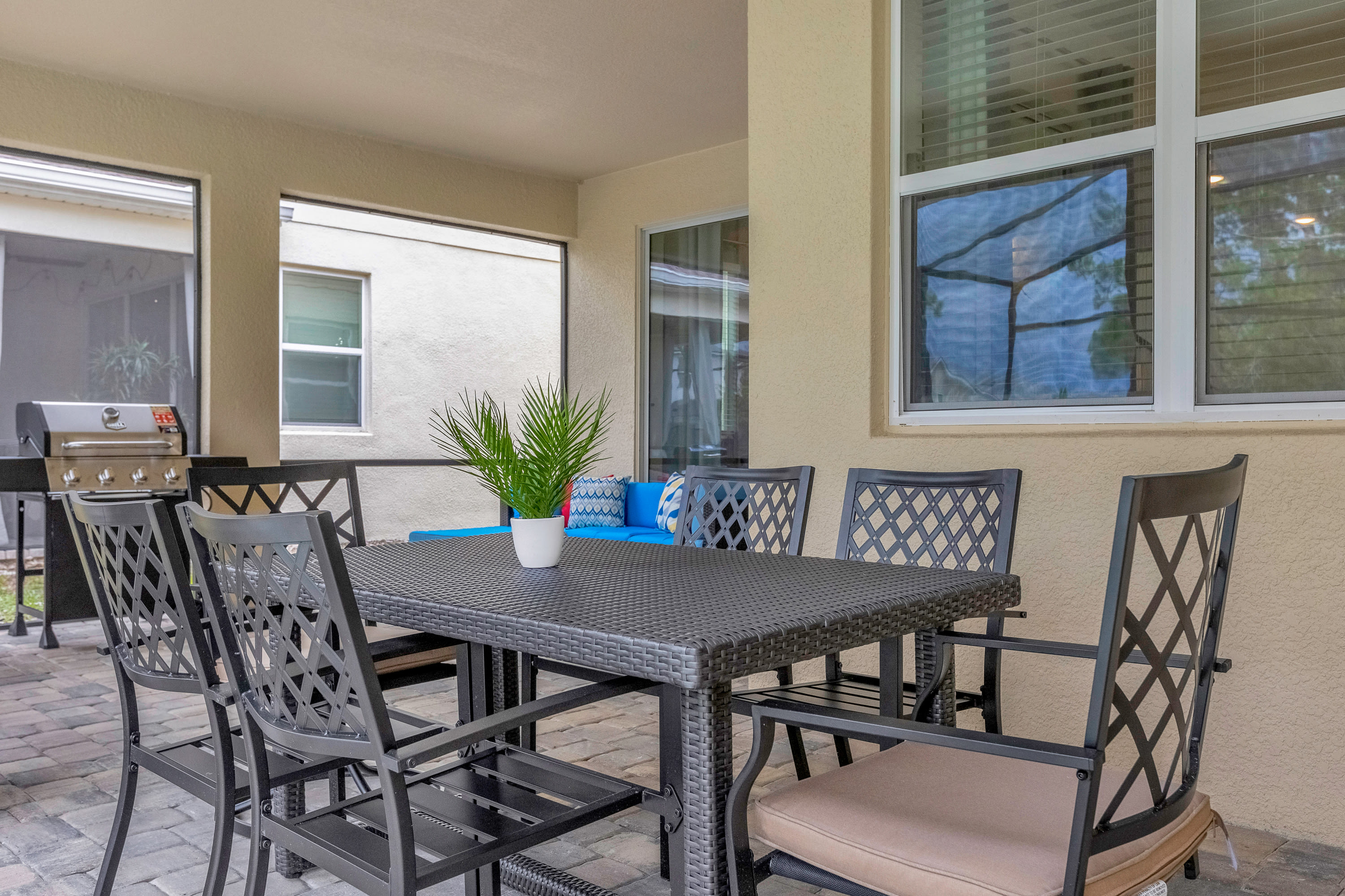 A covered outdoor patio features a dark wicker dining table with six chairs, a potted plant centerpiece, and a BBQ grill visible in the background, all on a paved surface next to large windows.