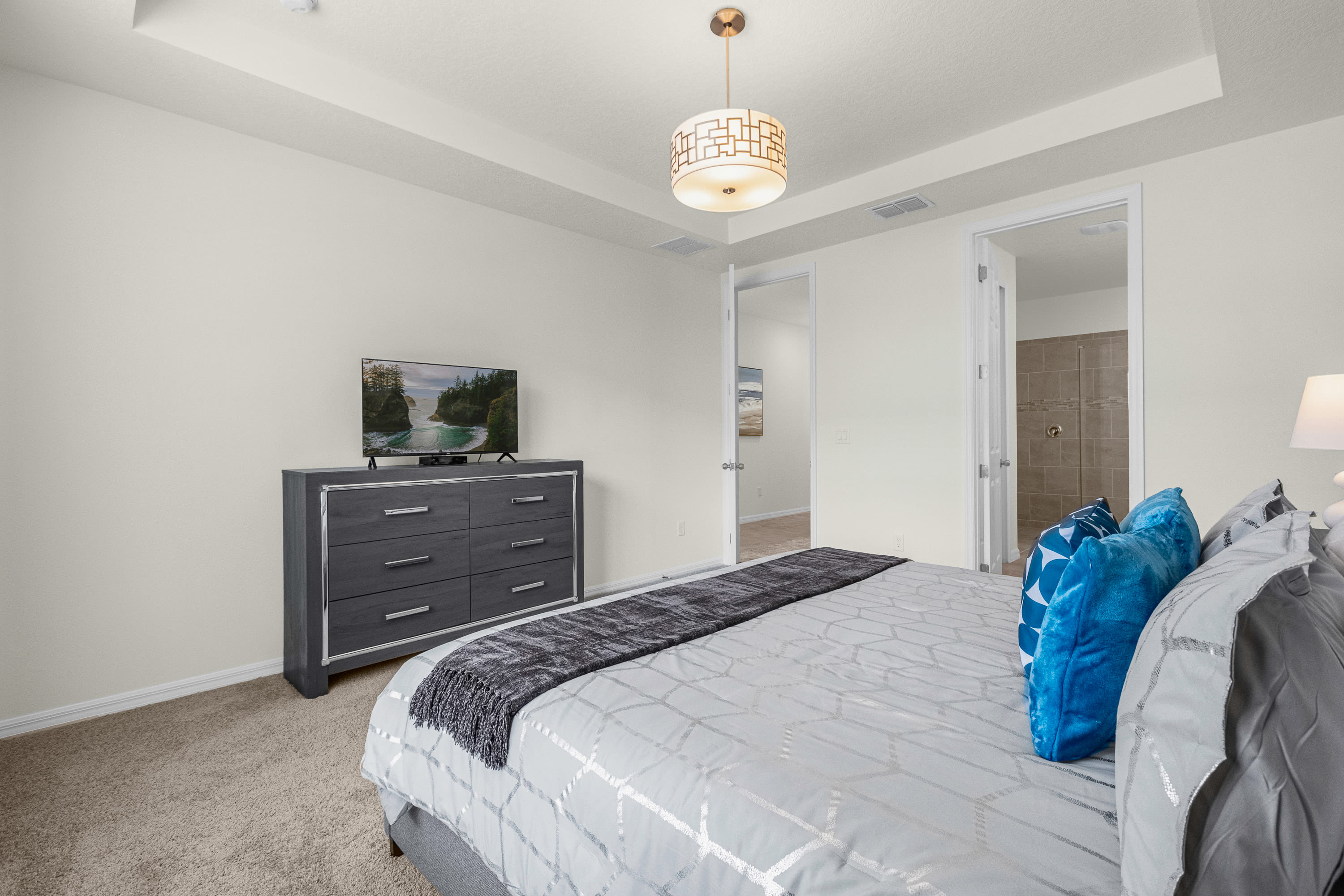 View of the master bedroom showing a flat-screen TV on a gray dresser, with doors leading to the en-suite bathroom and hallway, maintaining the room's neutral and blue color scheme.